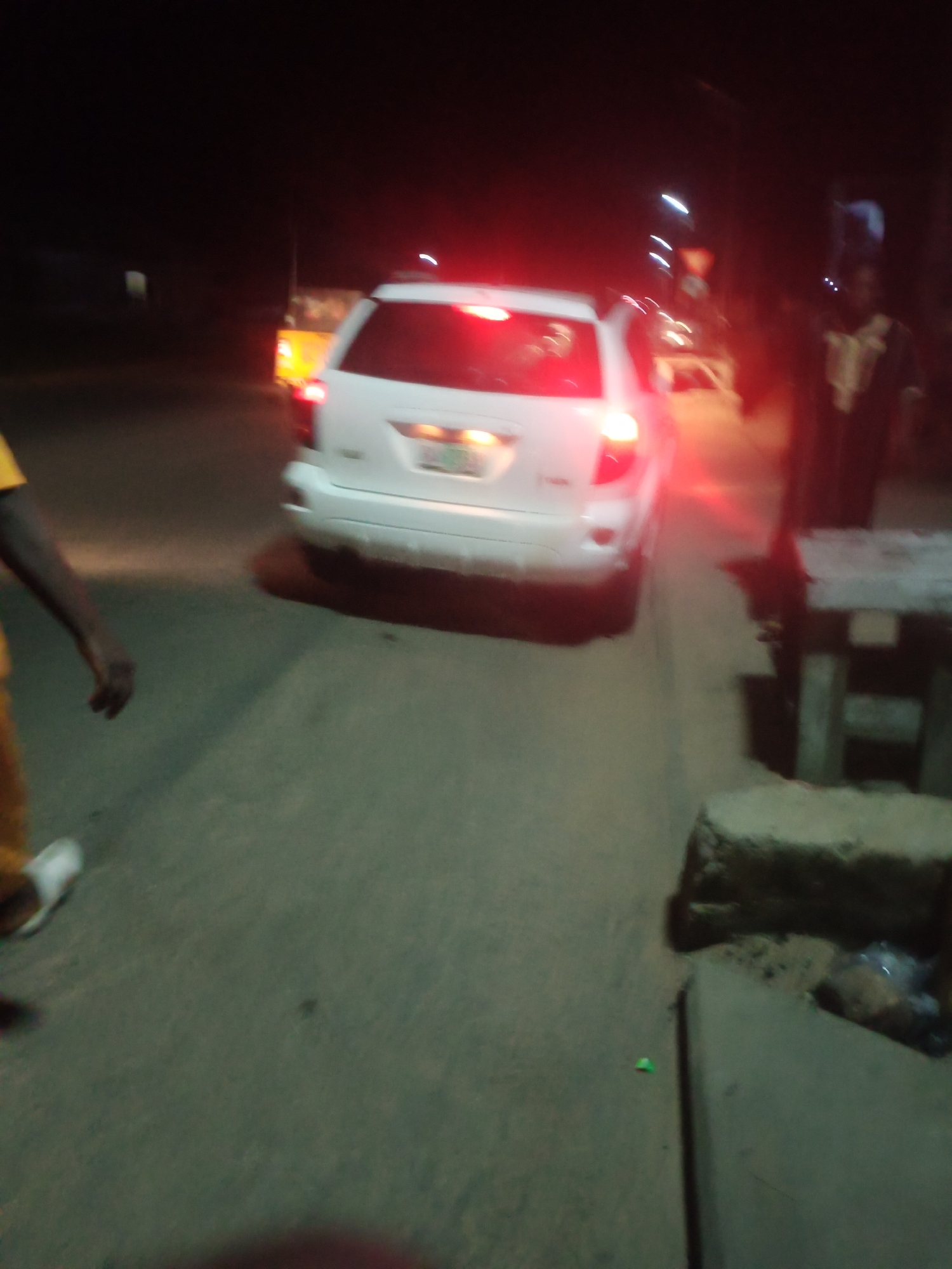 The image depicts a nighttime scene on a paved road, likely in Jalingo, Nigeria. A white SUV is the central subject, its taillights illuminated and casting a red glow. To the left, a person wearing a yellow garment and white footwear is partially visible, with their arm extended. To the right, another individual in dark clothing with a light-colored patterned garment is standing. In the background, blurred lights suggest other vehicles and structures, including what appears to be a yellow vehicle. The environment is characterized by a dimly lit road surface and roadside elements, including a concrete structure and what seems to be a wooden bench. The overall impression is that of a busy or traffic-filled street at night.