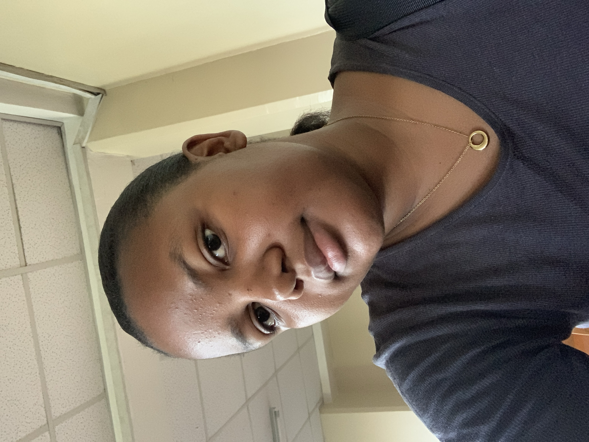 A medium close-up, portrait-oriented image (originally rotated 90 degrees) features a person with dark skin looking directly into the camera. The individual, appearing to be female, has dark hair neatly pulled back from her face. She wears a dark, possibly charcoal grey or black, short-sleeved top. A thin gold chain necklace with a small, simple gold ring pendant is visible around her neck. Her expression is calm and direct, with a subtle, soft smile, and her eyes engage with the viewer. There is a slight sheen on her forehead and nose, suggesting natural skin or the effect of indoor lighting.
The background indicates an indoor setting. To the left, a light-colored wall (cream or light yellow) and a portion of a white or light grey window or door frame are visible. Above and to the right, a white ceiling with a grid pattern of acoustic tiles can be seen. The overall lighting is consistent with ambient indoor illumination. The image appears to be a selfie. No text is visible in the frame. The location context for this image is Dar es-Salaam, Tanzania.