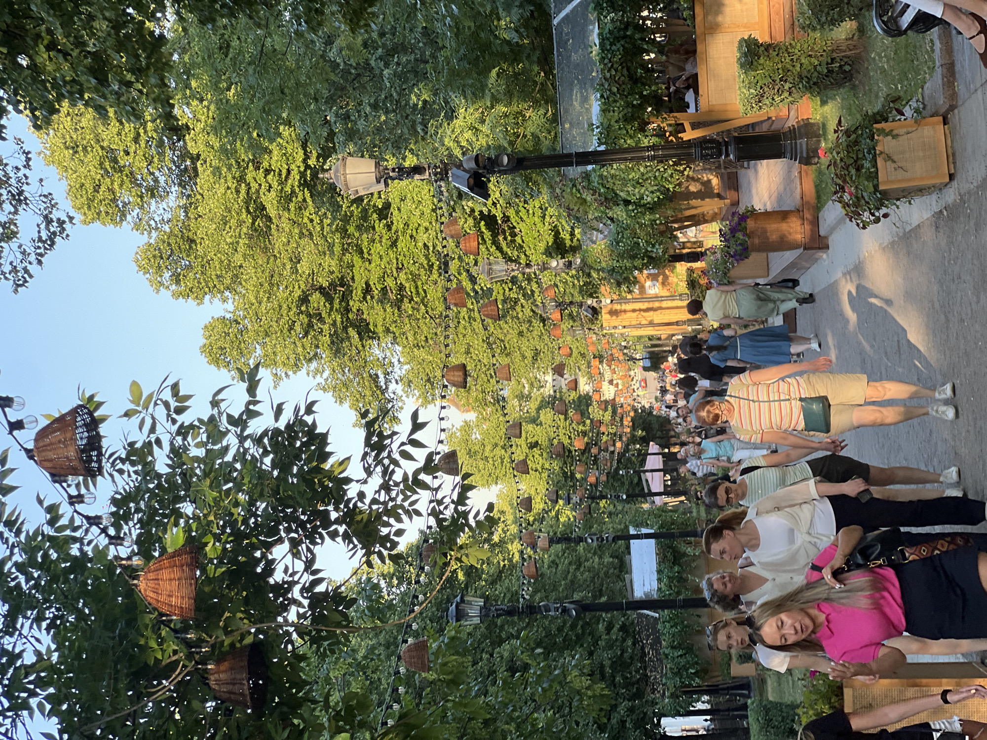 The image depicts a bustling pedestrian street in Moscow, Russia, likely during a warm season. The street is lined with mature trees providing ample shade, with string lights featuring woven basket-like lampshades and bare bulbs crisscrossing overhead. On the right side of the street, there are outdoor seating areas with tables and chairs, some covered with awnings, suggesting cafes or restaurants. People are walking in both directions along the paved pathway. A prominent black lamppost with a vintage design stands tall on the right. The overall atmosphere is lively and suggests a popular recreational area within the city.