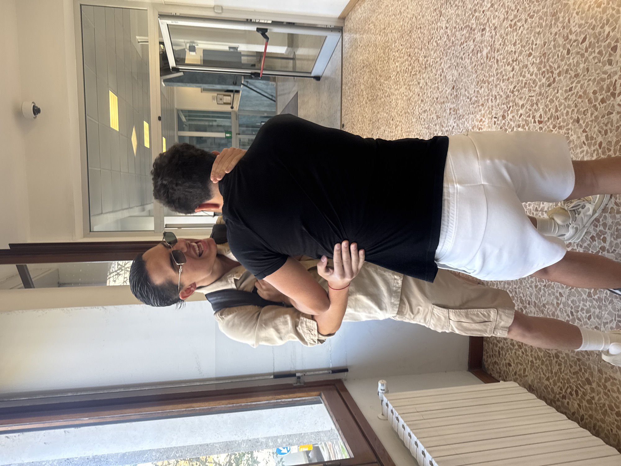 The image captures two young men engaged in a playful embrace or interaction within an indoor hallway, likely in a public building or office setting in Milan, Italy. The photo is taken from a slightly low angle, showing them in motion.
One man, with his back mostly to the camera, has dark, curly hair and is wearing a black t-shirt and white shorts with white sneakers and socks. He has his arms wrapped around the other man's lower back.
The second man, facing the camera, has neatly styled dark hair and is wearing dark sunglasses. He is smiling widely, mouth open in laughter, and wears a tan collared shirt or light jacket over a dark top, with tan cargo shorts and a backpack slung over one shoulder. His arms are around the first man's neck or upper back. A small red string bracelet is visible on his right wrist.
The setting is a well-lit corridor with a light-colored, patterned terrazzo-style floor. White walls line the hallway, and large glass windows or partitions are visible, reflecting rectangular interior lights. A white security camera is mounted on the left wall. In the background, a glass door at the end of the hallway and what appears to be a white radiator on the right side are discernible. The bright lighting suggests it is daytime. No discernible text is visible. The mood appears lighthearted and joyful.