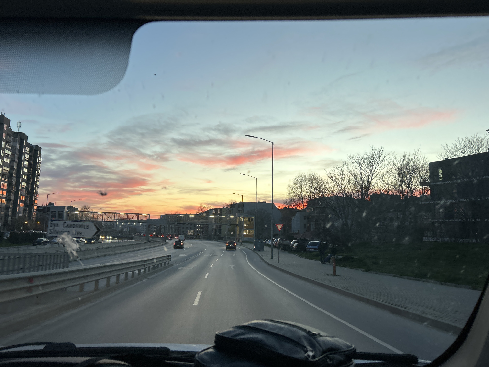 The image is taken from inside a vehicle, looking out through the windshield. A road stretches ahead, with multiple lanes and other vehicles. The road is flanked by a barrier on the left and a sidewalk with parked cars on the right. Street lights illuminate the scene. Buildings are visible on both sides of the road. The sky is a mix of colors, indicating a sunset or sunrise. A road sign is visible on the left which includes the words "бул. Сливница" which is "Bul. Slivnitsa" in the Cyrillic alphabet. The location is likely in or near Varna, Bulgaria, as "бул. Сливница" is a street in Varna.