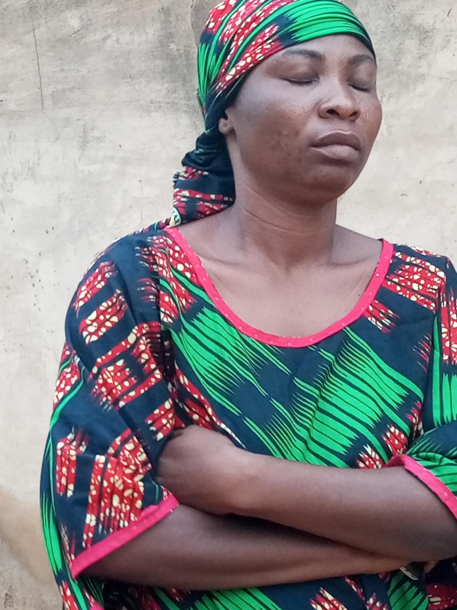 In the city of Tempure, Nigeria, a woman stands with her eyes closed, her arms crossed in front of her. She wears a vibrantly patterned headscarf and a matching dress, both adorned with green, red, and black designs. The background is a rough, textured wall, likely made of concrete or stucco, with a neutral, beige tone. The lighting appears to be natural daylight, casting subtle shadows on her face and the wall.