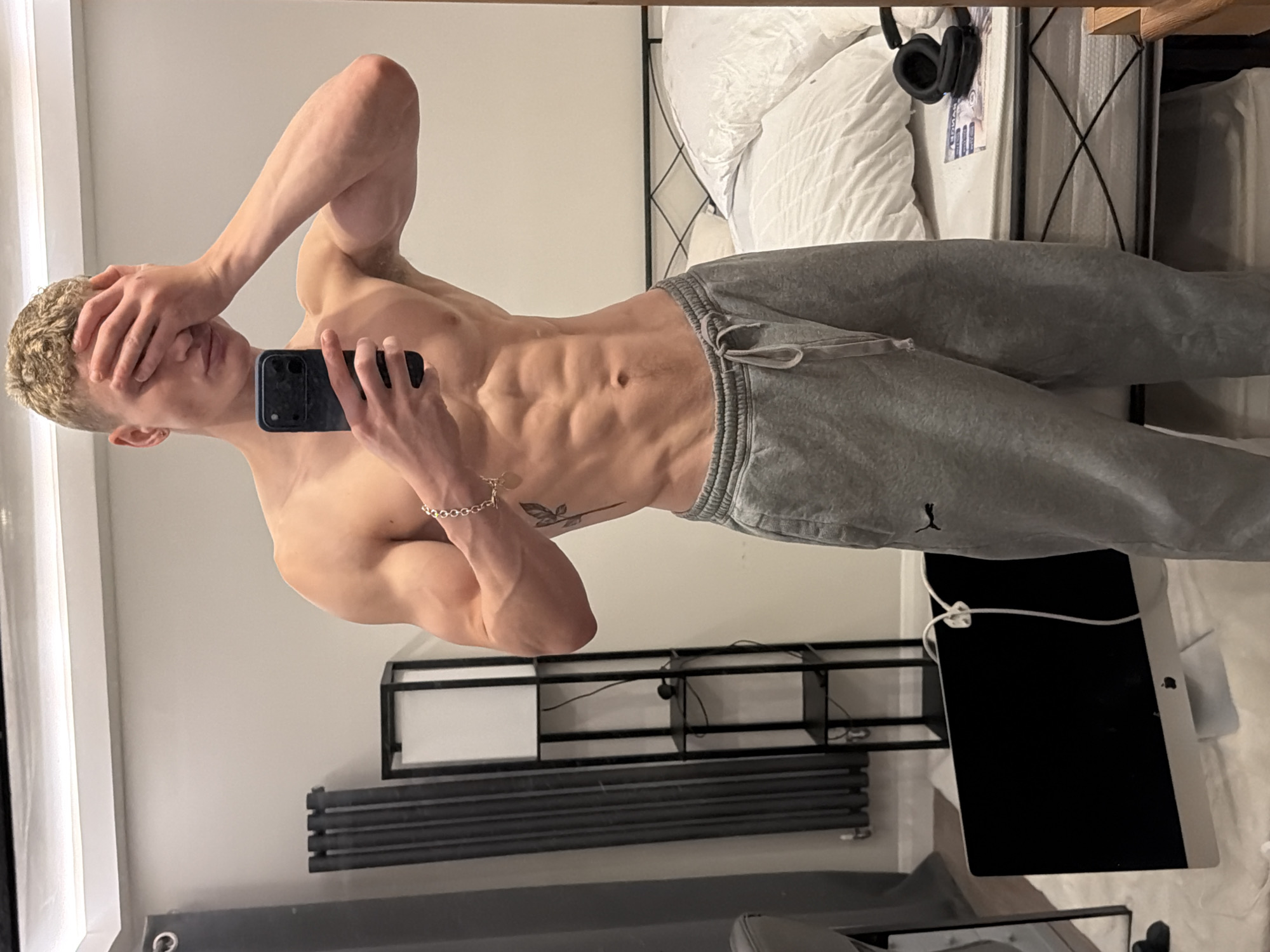 A male figure with a muscular physique is positioned indoors, likely in a bedroom environment. He is wearing grey sweatpants and holding a black smartphone, obscuring part of his face. His other hand is also covering his face. His skin is pale, and his hair is short and blond. A silver bracelet adorns his wrist, and a small tattoo is visible on his torso. The bedroom contains a bed with white bedding, black headphones resting on the bed, and an iMac screen. Behind the figure are a black metal shelf unit and a dark radiator. The scene suggests a private residential setting. The location in London, United Kingdom cannot be determined from the provided image.