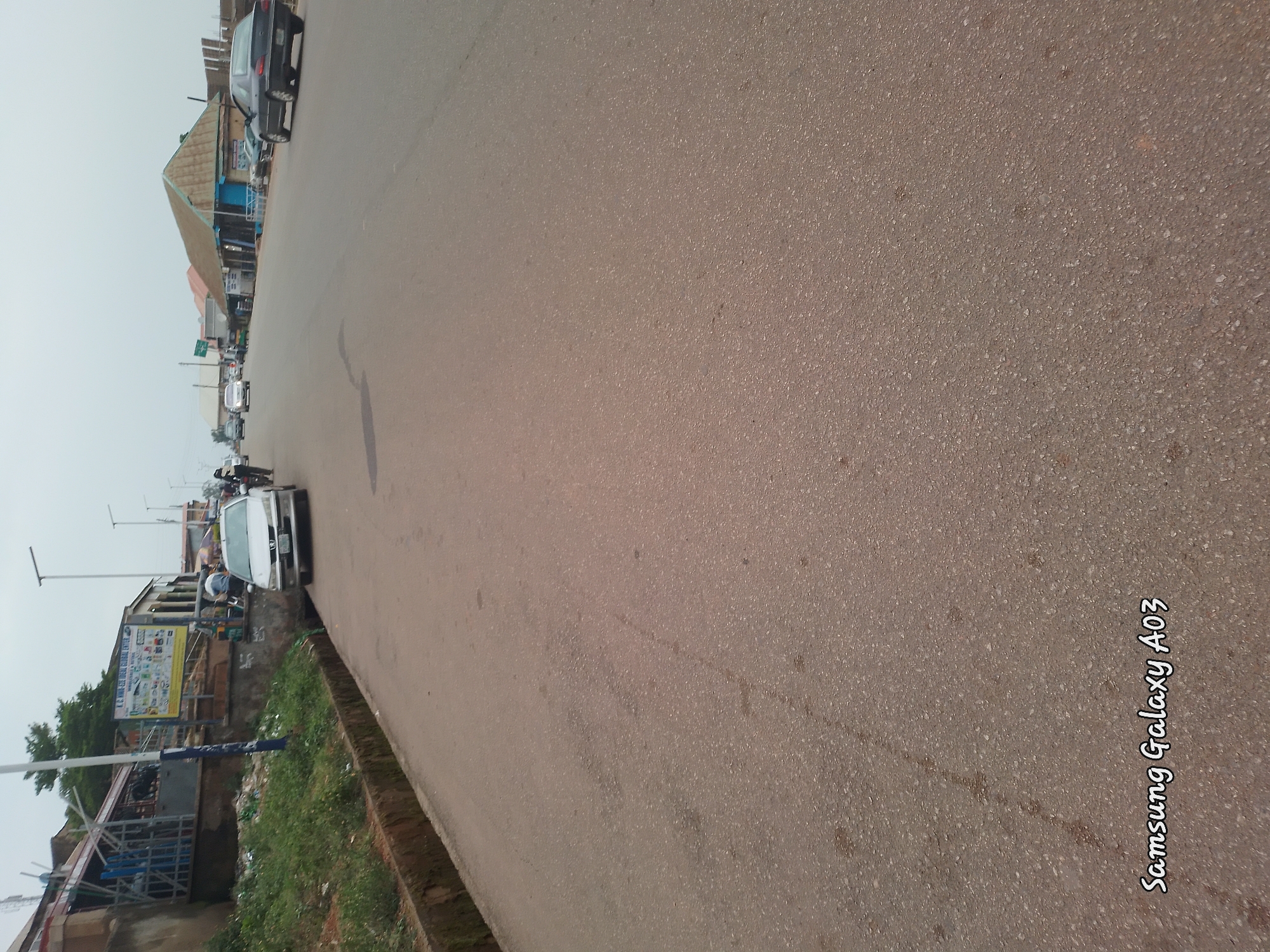 The image depicts a street scene in Jos, Nigeria, under an overcast sky. The foreground is dominated by a paved road with a textured surface. To the left, a raised concrete drainage channel runs alongside the road, filled with some debris and overgrown grass. A white car is parked on the right side of the road, facing away from the viewer, and further down the street, another car is visible.
Along the left side of the road, a row of shops and buildings line the street. One building has a blue and white facade with metal bars on the windows. A large, rectangular sign with text and images is prominently displayed on this building. A person on a motorcycle is visible near this sign.
Further down the street, more shops with corrugated metal roofs are visible. The road appears to be a main thoroughfare, with several cars and a motorcycle traveling in the same direction as the viewer. The environment suggests an urban setting within Jos, likely a commercial area due to the presence of numerous shops. The overall impression is of a typical day in the city, with moderate activity on the street. The text "Samsung Galaxy A03" is watermarked in the lower left corner of the image.