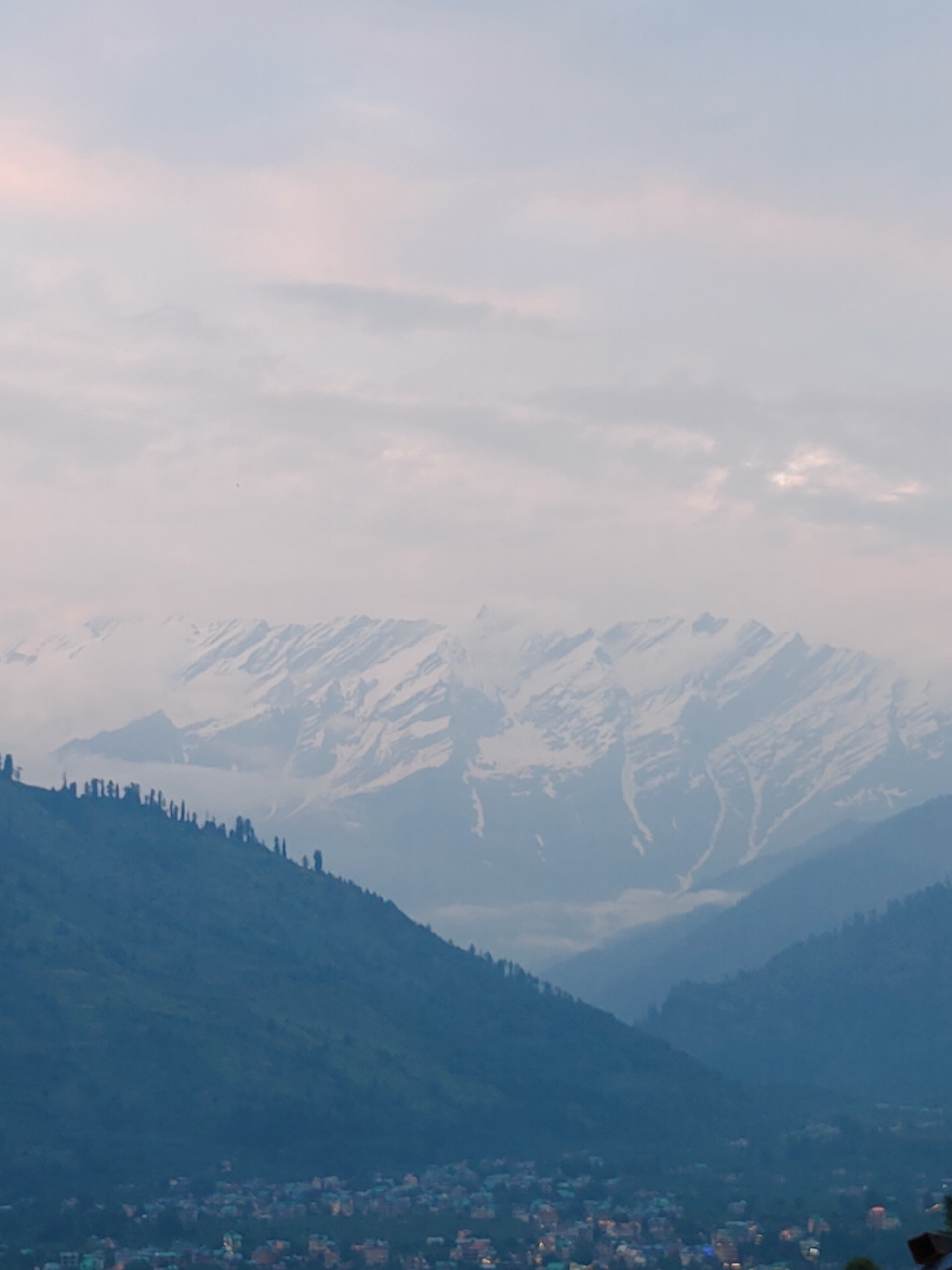 The image shows a panoramic view of the town of Vashisht, India, nestled in a valley with a backdrop of snow-capped mountains. The foreground features a densely populated area with numerous buildings, possibly residential or commercial, characterized by their varied architecture and illuminated windows. The mid-ground is dominated by a steep, tree-covered mountain slope that descends into the valley. The trees appear to be coniferous, with pointed tops, creating a dark, textured silhouette against the lighter sky. In the background, a majestic mountain range rises, its peaks covered in snow and partially obscured by atmospheric haze and clouds. The sky itself is overcast with soft, diffused light, suggesting it might be early morning or late afternoon. The overall scene evokes a sense of natural beauty and a remote, mountainous settlement.