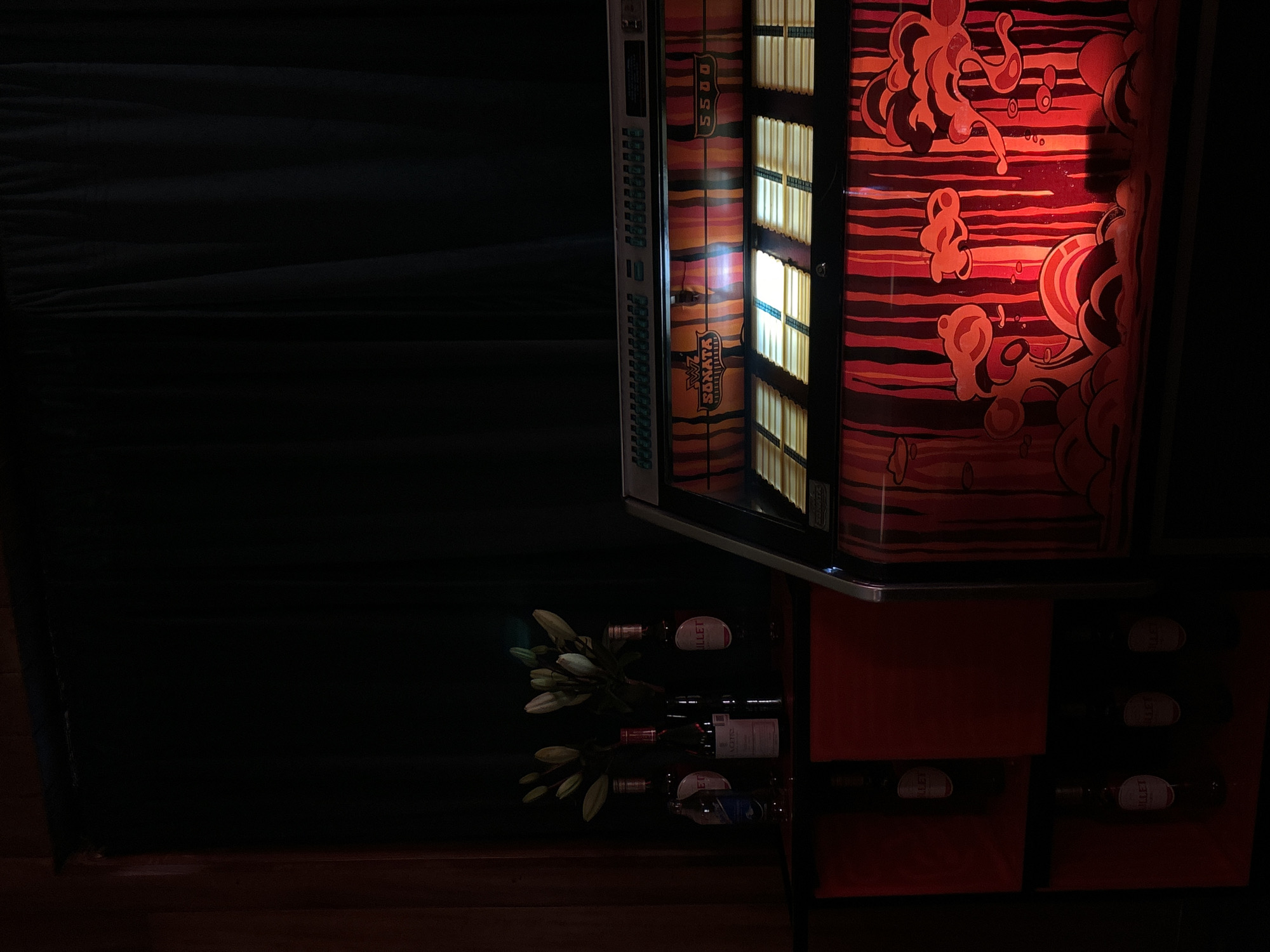 A partially lit vintage jukebox with decorative artwork stands in a dark room. Below it, there is a red cabinet housing bottles of wine. Some white flowers are placed next to the wine bottles. The room is dimly lit.