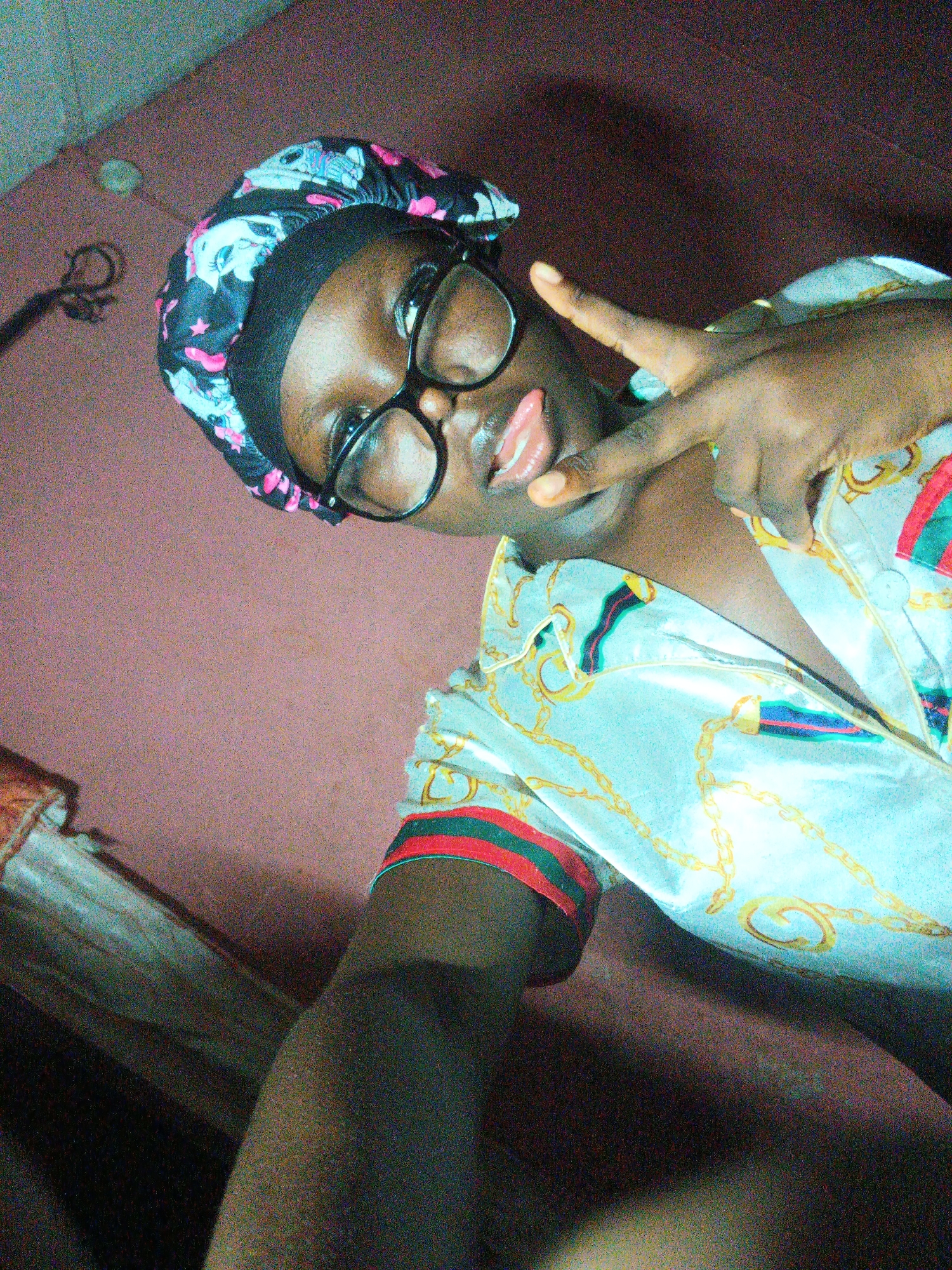 A close-up selfie shows a person, likely a young woman, posing indoors. She has dark skin and is wearing black-framed glasses, which rest on her nose. On her head, she wears a black bonnet with a vibrant pattern featuring pink and light blue elements, including what appear to be cartoon character-like figures and stars. Her tongue is playfully sticking out, and her left hand is raised, forming a peace sign with two fingers.
She is wearing a light-colored, possibly satin, shirt or pajama top with a patterned design. The shirt features prominent gold chain motifs and has red and green striped cuffs or trim details. The image quality is grainy, suggesting it was taken in low light conditions, possibly with a mobile phone.
The background is out of focus, showing a reddish-brown floor or carpet and some indistinct dark objects. The photo is tilted, indicating a casual, handheld shot. There is no visible text in the image. The overall impression is one of a relaxed and playful moment.