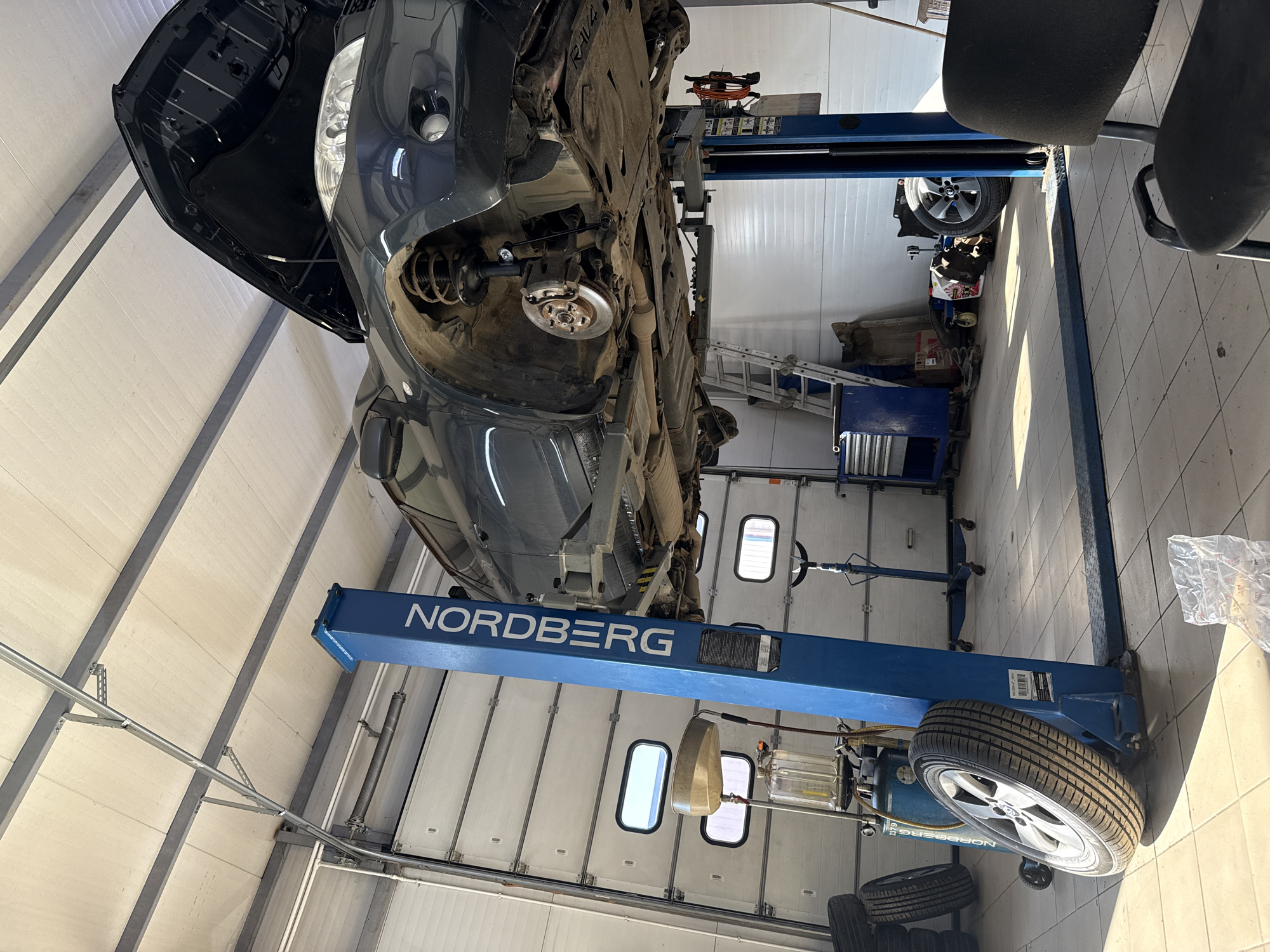 A dark-colored vehicle is elevated on a blue two-post car lift in a garage. The vehicle's hood is open, and the front passenger-side wheel is missing, revealing the suspension and brake components. The garage features white roll-up doors in the background, and various tools and equipment are visible, including stacked tires, an oil drainer, and a toolbox. A ladder leans against a wall, and a chair is situated in the foreground. The scene appears to be a car repair shop.