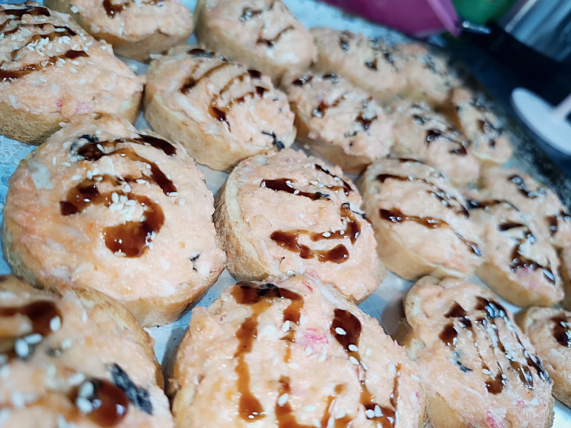 The image shows a close-up of a tray filled with multiple pieces of what appear to be Japanese-inspired appetizers. Each piece consists of a slice of toasted bread topped with a pinkish-orange spread, likely a spicy tuna or salmon mixture. A dark sauce, possibly eel sauce or a similar glaze, is drizzled over the top, and sesame seeds are sprinkled as a garnish. The location is unknown, and the query for Chaikovsky, Russia, is not applicable. No people or actions are visible.