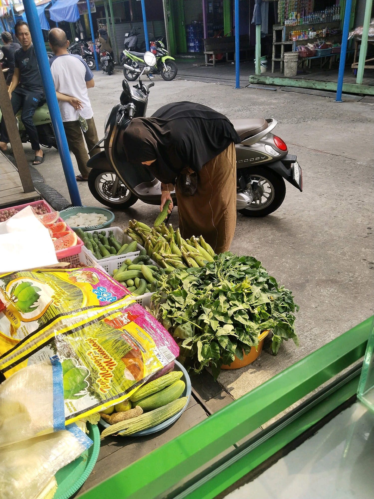 An outdoor street market scene in Muara Muntai Ulu, Indonesia, depicts a person in a black headscarf and brown hijab-like garment bending over to examine a pile of leafy green vegetables in a yellow plastic basin. They are reaching out with their right hand, holding a green cucumber. In the foreground, to the left, are several plastic bags with colorful packaging, one featuring an image of coconuts and text in Indonesian. Behind these bags, to the left and center, are plastic crates filled with various vegetables, including more cucumbers, shallots, and what appear to be slices of watermelon in a pink crate. Behind the person, a motor scooter is parked. Further in the background, other people are visible near parked motorcycles and market stalls with shelves displaying goods. The ground is paved with concrete, and blue and green poles are present, suggesting a market structure.