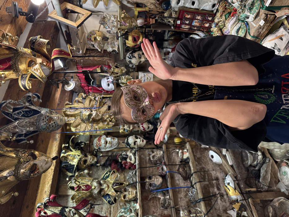 An indoor photograph depicts a young adult individual wearing an ornate Venetian half-mask within a mask shop in Venice, Italy. The individual, positioned centrally, wears a light pink and gold Colombina-style mask covering her eyes and nose. Her hands are raised symmetrically to shoulder height, palms open, and her lips are puckered. She wears a black t-shirt over a dark blue apron featuring "VENEZIA" text and a laurel wreath, along with a long chain necklace. A silver ring is on her left ring finger. The background is densely populated with a wide variety of Venetian Carnival masks, including full-face, half-face, and jester designs, displayed on walls, shelves, and hanging from the ceiling. Wooden beams are visible overhead, and artificial lighting illuminates the scene. Unfinished mask forms are visible on lower shelves to the left, and a wooden counter with additional masks is present in the lower right.