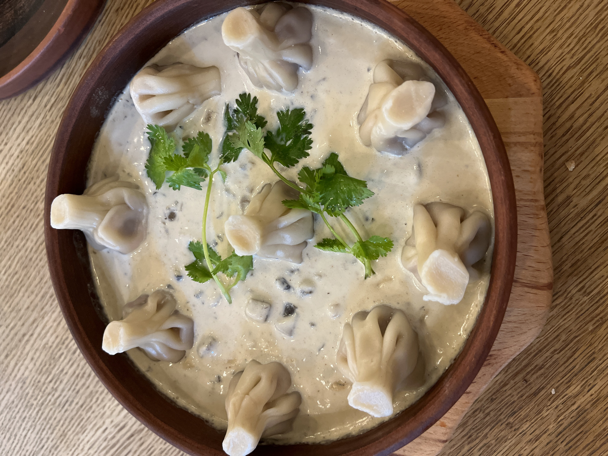A close-up, overhead shot shows a rustic, dark brown ceramic bowl filled with a creamy mushroom soup. Submerged in the soup are eight khinkali dumplings, each with a distinct pleated top knot. A few sprigs of fresh cilantro are scattered on top, adding a touch of green to the dish. The bowl rests on a wooden serving board, which is placed on a light-colored wooden table. The lighting is natural and soft, highlighting the textures of the food and the wood. The scene is set in a restaurant in Sochi, Russia.