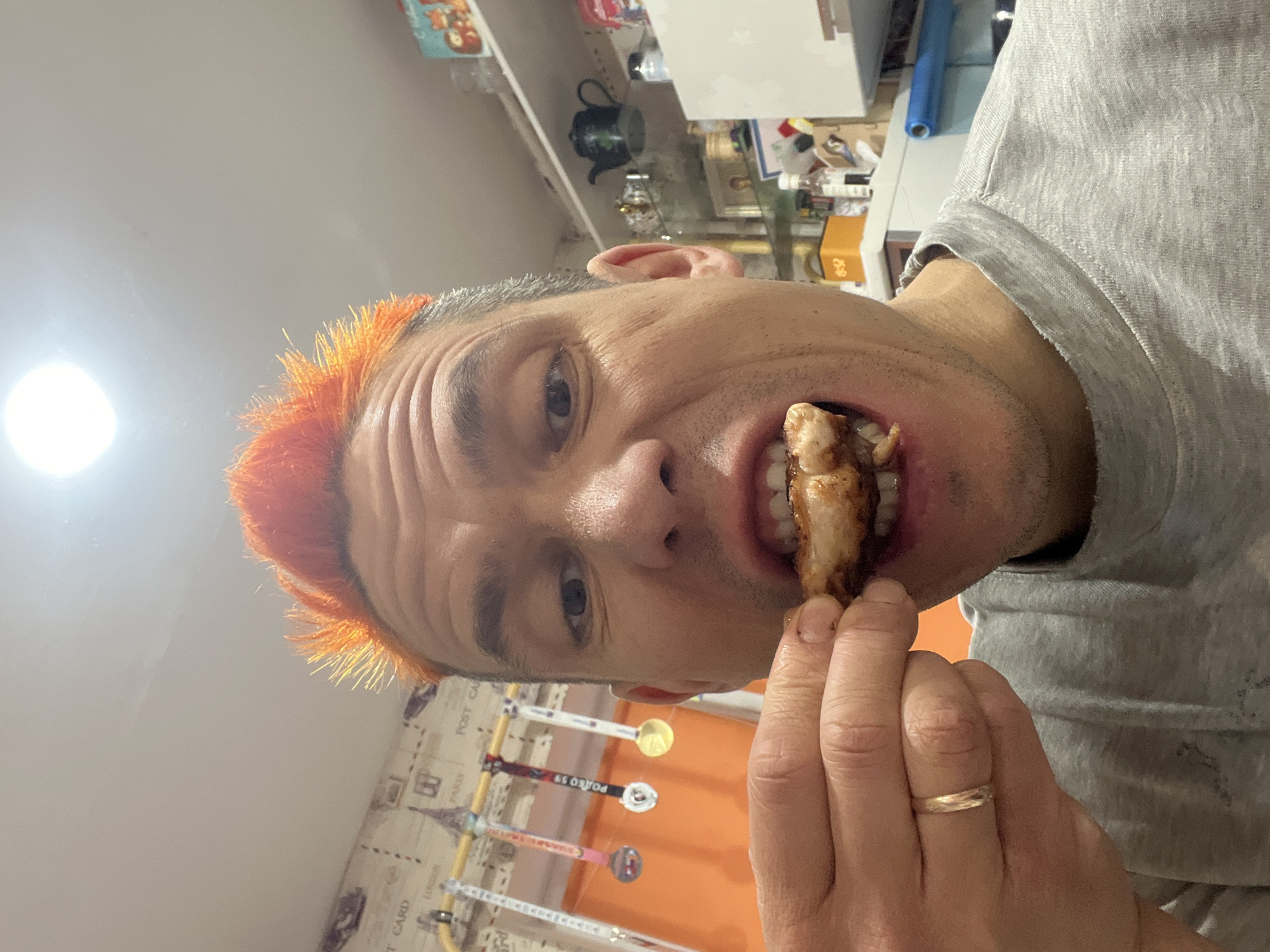 A close-up, slightly rotated image features a person with short, spiky, orange-colored hair. The person is light-skinned with visible facial wrinkles. The person is in the act of eating a piece of what appears to be cooked meat, holding it close to their mouth with their right hand, in which the ring finger displays a simple gold band. The person is wearing a gray t-shirt.
In the background, a wall decorated with a pattern including images of Eiffel towers and the words "POST CARD" and "PARIS" can be seen. Four medals are displayed on the wall. On the left side of the wall, there is a long orange thing. A glimpse of shelves stocked with various items including a black teapot and a gold-colored box is visible, alongside a blue rolled-up object. The brightness is higher on the left side of the image, and the image appears to have been taken indoors.
The location of the picture is not possible to determine with the visible elements of the image. The city of Chaikovsky, Russia is not identifiable with the context of the image.
