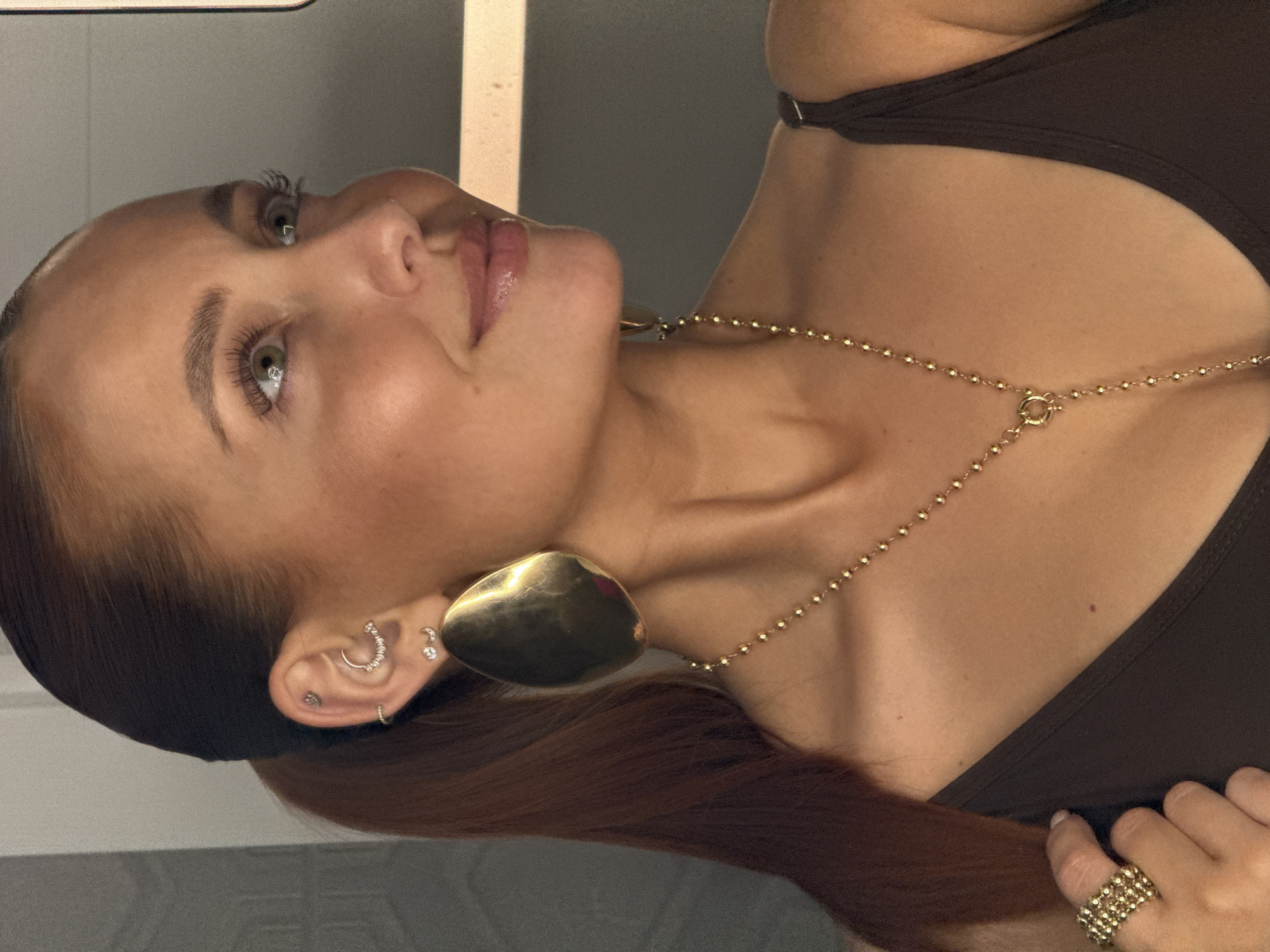 A woman is pictured from a low-angle perspective, wearing a brown tank top. She has light skin, light eyes, and dark brown hair, with some strands of red hair visible. She is wearing gold earrings in various styles on her left ear, including a large, heart-shaped earring and smaller studs. A gold necklace is around her neck. She is wearing a ring on her right hand. The background appears to be a room with a grey wall and a beige object. There is no information available to connect this image to Chaikovsky, Russia.