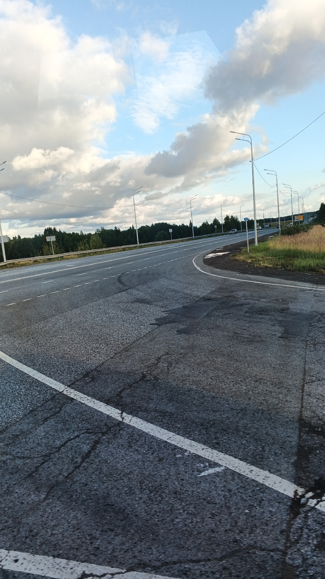 The image depicts a road intersection with several lanes, a wooded area in the background, and a cloudy sky. The road surface is asphalt, showing signs of wear and tear, with visible cracks and patches. White lane markings are present, including a solid white line on the left side of the frame. Streetlights line the side of the road, and some signs are visible in the distance. A few vehicles are driving on the road. The surrounding environment appears rural or semi-rural, with a forest bordering the road and fields to the right. The lighting suggests it is daytime, possibly late afternoon or early evening, given the cloud cover and the quality of light. There are no people visible in the image.