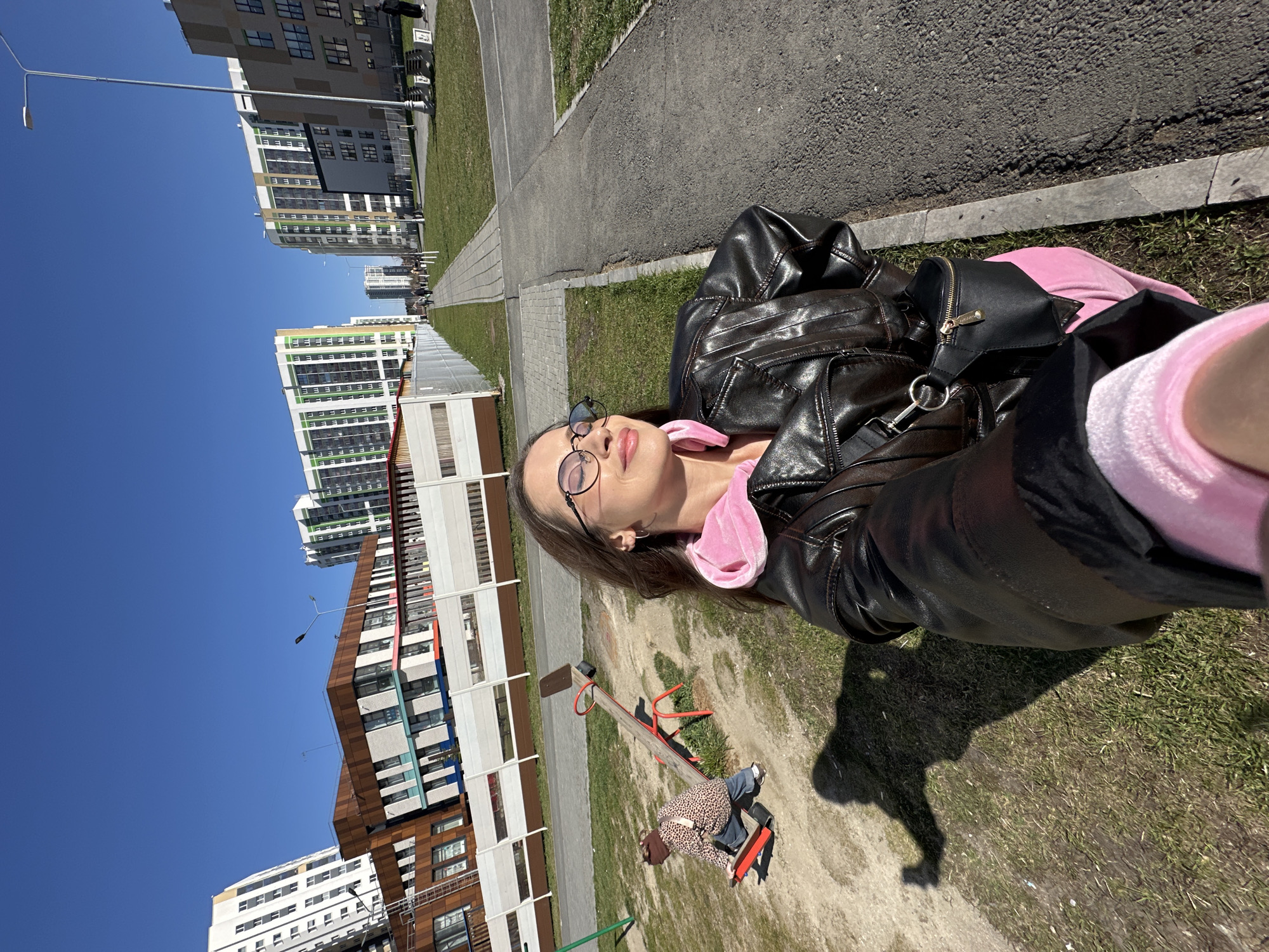 A woman in a pink hoodie and black leather jacket lies on the grass in Yekaterinburg, Russia, taking a selfie. She is wearing glasses. Behind her is a seesaw with a child riding on it. In the background are modern apartment buildings and a clear blue sky. A paved pathway runs alongside the grass.