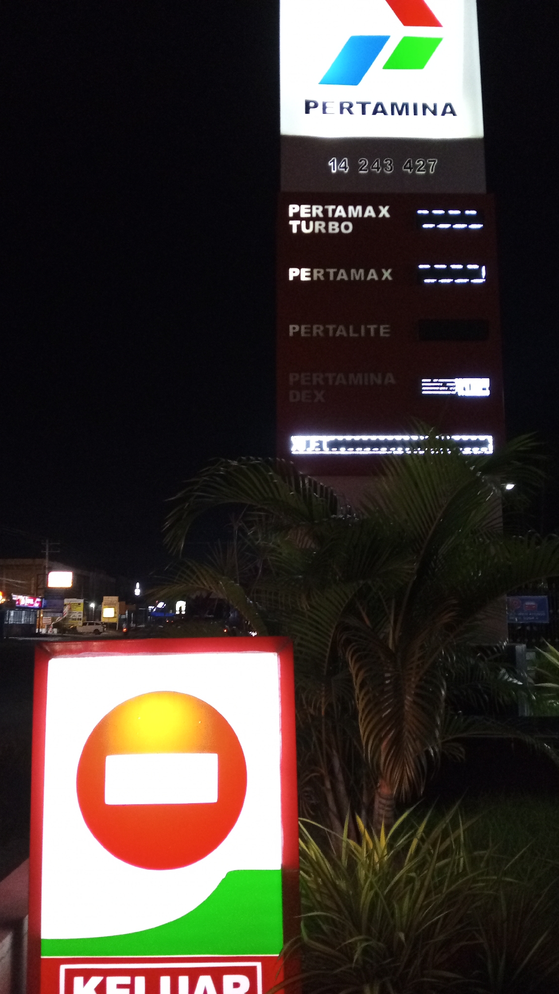 The image shows a night scene at a Pertamina gas station in Cotbadatunong, Indonesia. The main subject is a tall digital price board displaying various fuel types and prices. The Pertamina logo is visible at the top. Below the logo, the number "14 243 427" is displayed. The fuel types listed are PERTAMAX TURBO, PERTAMAX, PERTALITE, and PERTAMINA DEX, each with corresponding prices or symbols indicating pricing. The lower portion of the price board displays "JET." In the foreground, there is a prominent red and white sign with a red circle and a white horizontal bar, symbolizing a "no entry" or "stop" sign, with the word "KELUAR" (meaning "exit" in Indonesian) written below it. Behind the sign, a palm tree and other greenery are visible. In the background, other buildings and lights of the city can be seen. The overall scene is illuminated by artificial lights.
