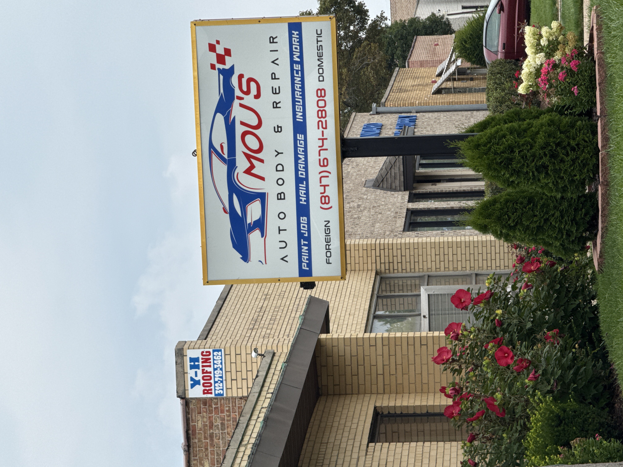 In Skokie, United States of America, a storefront displays two signs. The larger sign, a yellow rectangular billboard with a blue and white graphic of a car and the words "MOU'S AUTO BODY & REPAIR," advertises automotive services. Below this, in blue text, are the words "PAINT JOB," "HAIL DAMAGE," and "INSURANCE WORK," followed by "FOREIGN (847) 674-2808 DOMESTIC." A smaller, blue and white sign on the corner of the brick building reads "Y-H ROOFING" and a phone number "312-719-3462." The building itself is constructed of light brown brick. In front of the building, a large bush with vibrant red flowers is in bloom, along with manicured green bushes and trees. A red car is parked on the right side of the image, partially visible behind the greenery. The sky is overcast with light gray clouds.