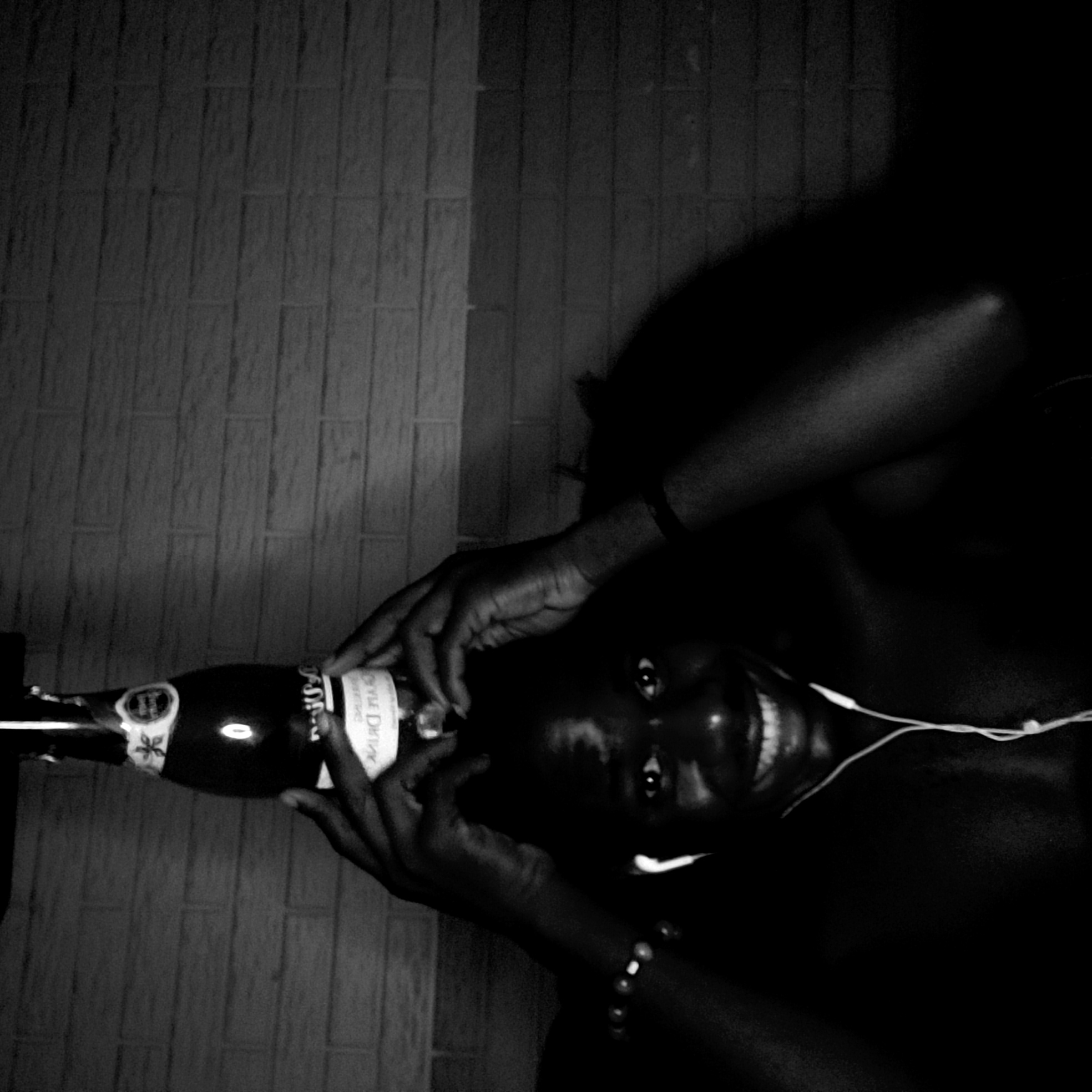 The black and white image shows a person lying down, holding a champagne bottle. The bottle is labeled "Rose Divine Sparkling". The person is wearing headphones. The background appears to be a wall with a brick or tile pattern. The location is Narayi, Nigeria.