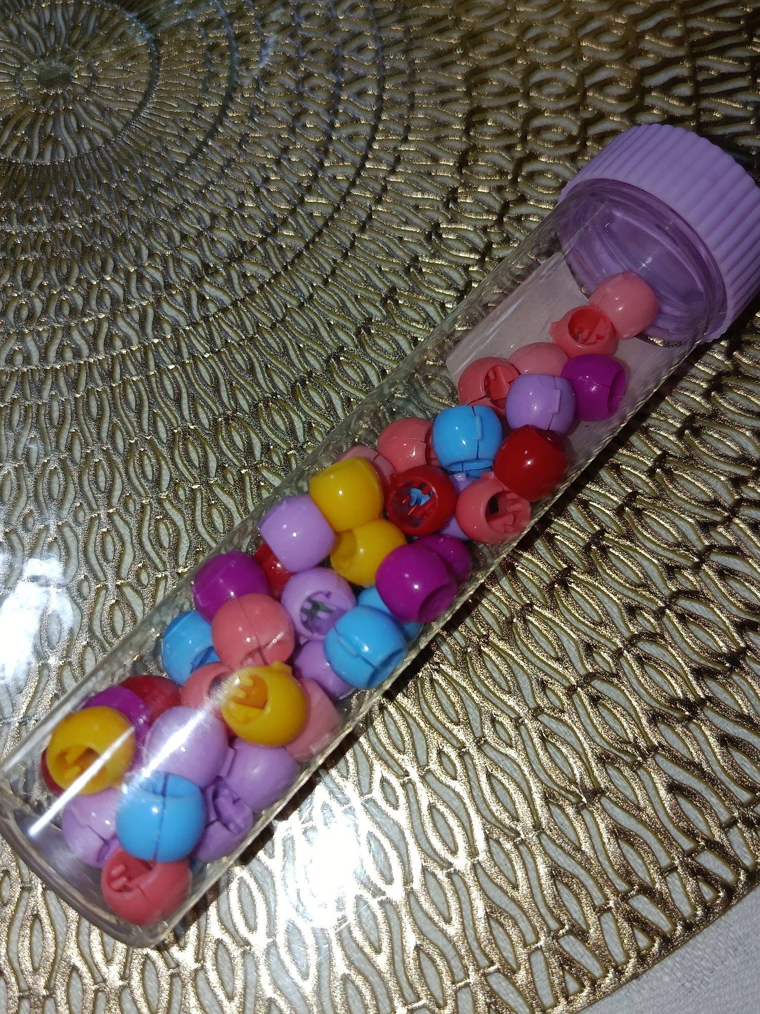 A clear plastic tube filled with small, colorful, hollow plastic beads is shown lying diagonally on a textured, golden placemat. The tube is capped with a lavender plastic lid. The beads are a mix of pink, purple, blue, yellow, and red. Some beads are partially open, revealing an inner cavity. The placemat has an intricate, woven pattern. The scene is illuminated by overhead lighting, creating reflections on the plastic tube and beads. The image does not contain any elements that would indicate a location in Tashkent, Uzbekistan.