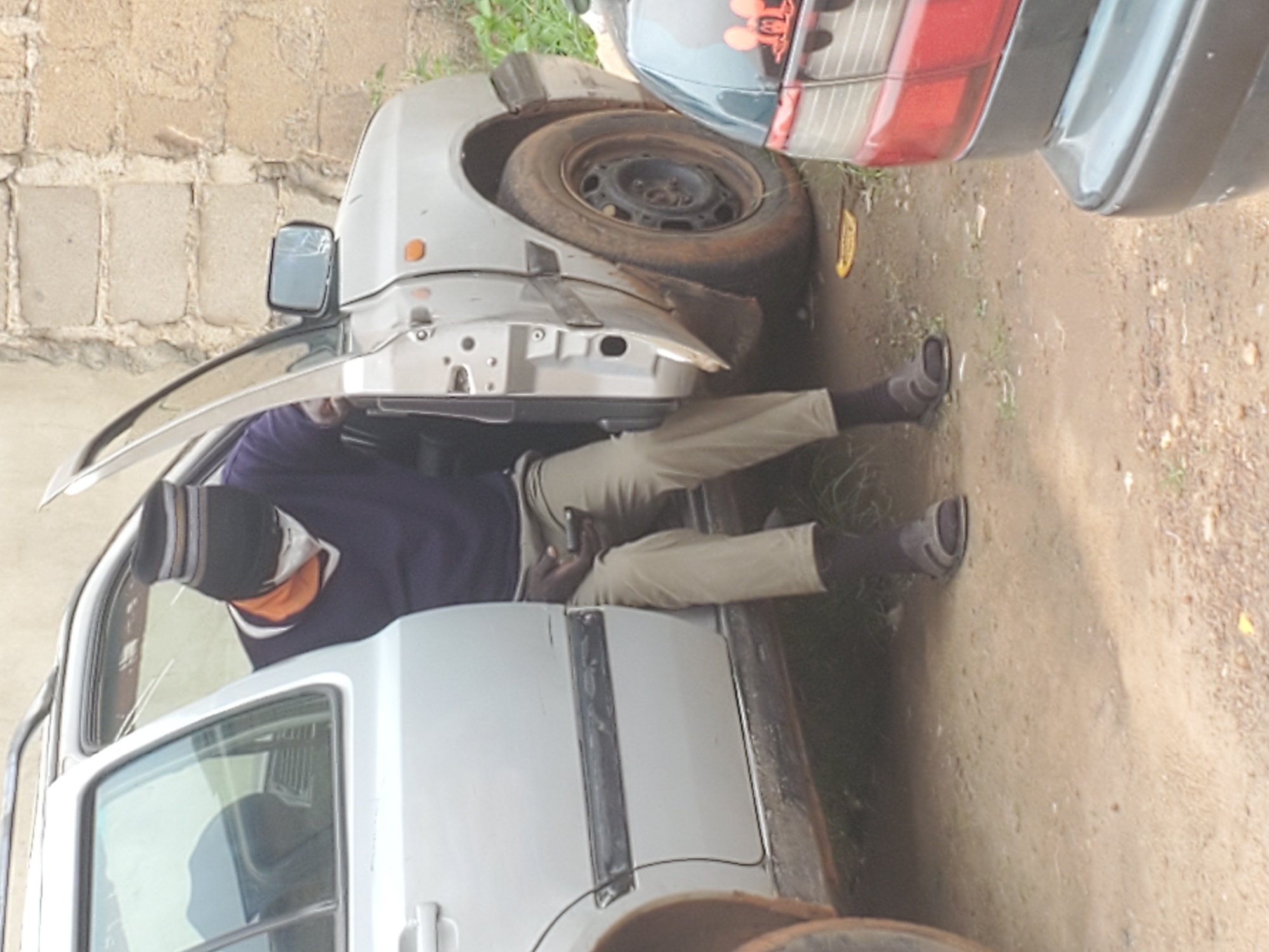 A person wearing a dark beanie with horizontal stripes, an orange t-shirt, a dark blue v-neck sweater, khaki pants, and dark sandals is sitting in the open driver's side of a silver station wagon. The person is looking down at a dark-colored cell phone held in their right hand and appears to be wearing a white face mask. The car has a roof rack and the driver's side door is fully open. The car is parked on a dirt road next to a light-colored brick wall. There is a second car, dark colored with red taillights, partially visible to the right of the station wagon. The environment is outdoors, likely in an urban or semi-urban area of Nigeria.