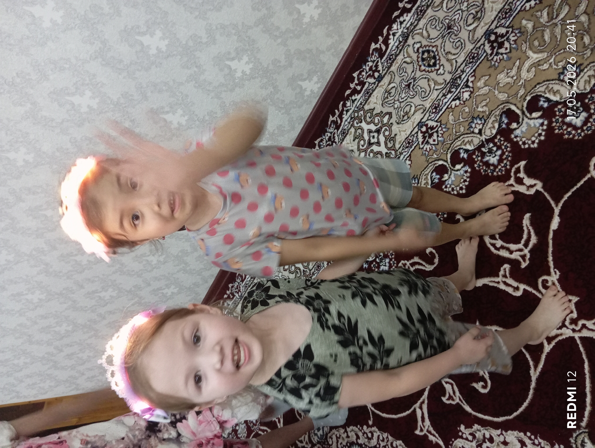 Two young girls are posing for a photo on an ornate rug. One girl is wearing a gray shirt with red polka dots and a white headband, waving her hand. The other girl is wearing a green floral dress and a tiara. The background includes a patterned wall. The photo was taken on May 17, 2026, at 20:41 with a Redmi 12 device. The location is likely indoors, possibly in a home. Bekabad, Uzbekistan is not explicitly visible or identifiable in the image.