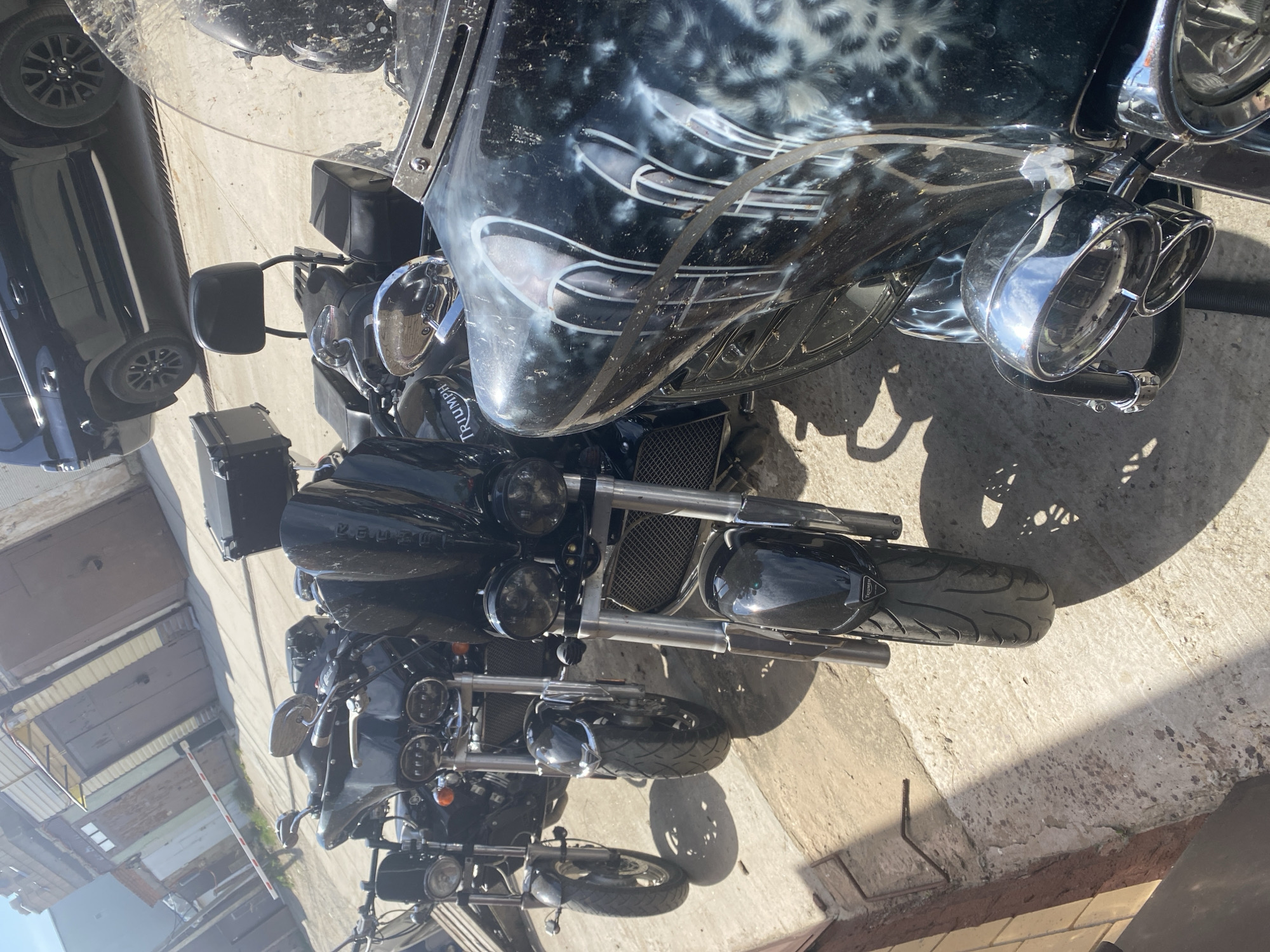 The image depicts a group of motorcycles parked on a concrete surface, likely a sidewalk or parking area. The motorcycles are predominantly black, with chrome accents. One motorcycle has a distinctive windscreen with a white and grey patterned design. A black car is partially visible on the left side of the image. The environment appears to be urban, with building facades and structures visible in the background. The location is identified as Chaikovsky, Russia. The image shows the motorcycles in a static, parked state. No people are visible in the scene. The composition is from an elevated perspective.