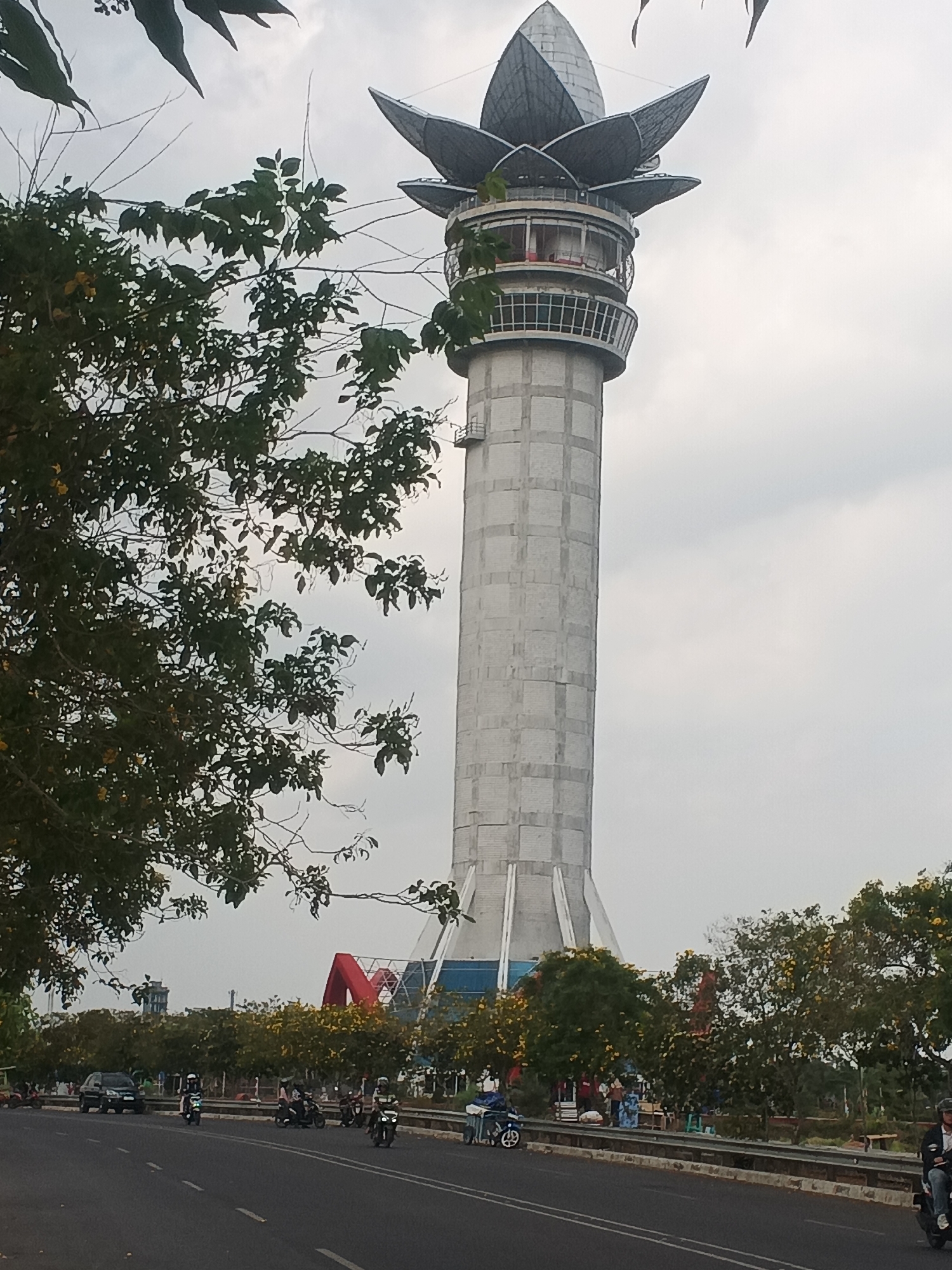The image shows the Taman Miniatur Koin Tower in Purwokerto, Indonesia. The tower, which resembles a blooming lotus flower, is tall and cylindrical with a unique architectural design at its apex. In the foreground, a road with traffic, including motorcycles and a car, is visible. Several trees with green foliage and some yellow flowers line the roadside, partially obscuring the view of the tower and other structures in the background. There are people standing and walking on the sidewalk near the trees, and some are gathered around food stalls. The sky is overcast.