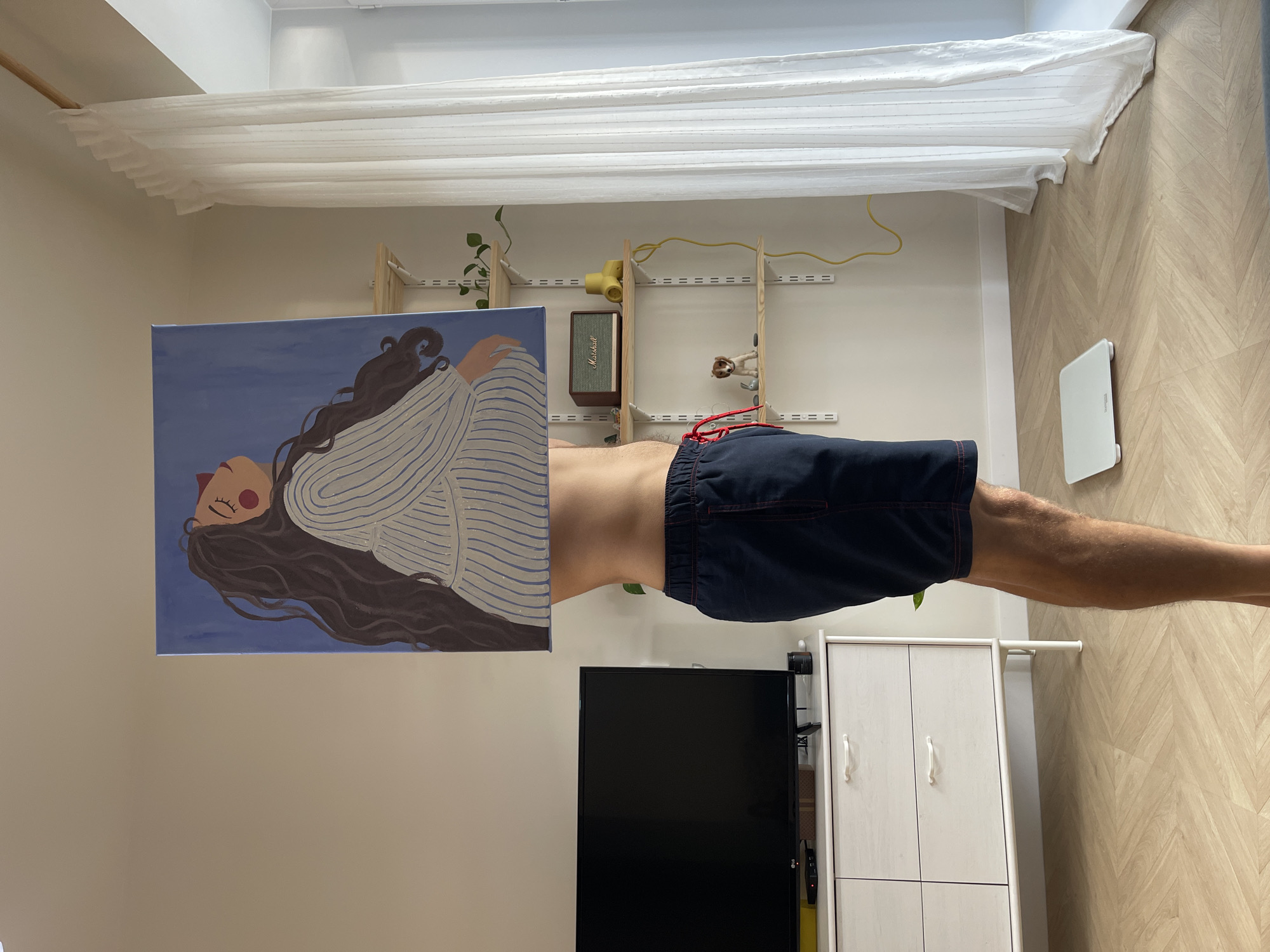 A man in dark blue shorts is levitating horizontally in a room. A painting of a woman is levitating beside him. The room contains a white cabinet, a black television, a wooden shelving unit, and a white scale. A white curtain is near the ceiling. The walls are beige and the floor is wooden.