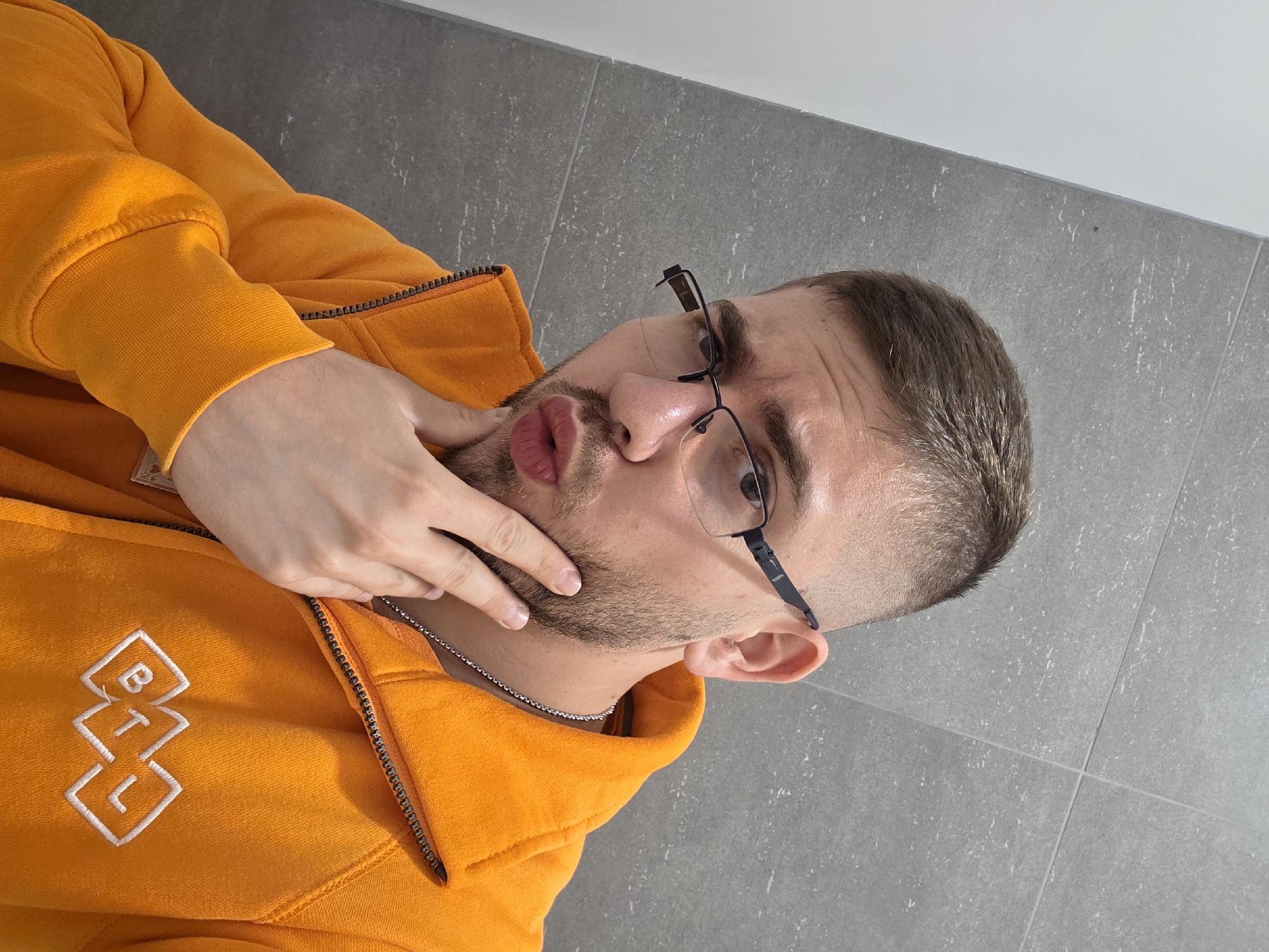 A man with short brown hair, a light beard, and glasses is posing for a selfie. He is wearing an orange hoodie with a white logo on the left side. His left hand is touching his chin. He's wearing a silver chain. The background is a grey tiled wall. The location cannot be determined to be Plovdiv, Bulgaria, based on the visual information provided.