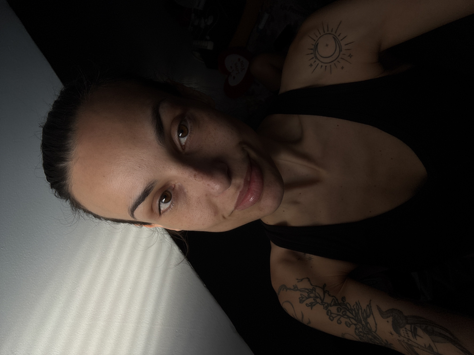 A woman is pictured in a close-up, angled shot. She is fair-skinned, with dark hair pulled back, and light brown eyes. Freckles are visible on her face. She is smiling slightly. She is wearing a black tank top. Tattoos are visible on her upper arms: one on her right shoulder depicting a sun and moon, and another on her left arm featuring a bird and floral designs. Light is entering from the left side of the frame, casting shadows on the adjacent wall. The backdrop appears to be a dimly lit interior. There is no information to indicate the image was taken in Pleven, Bulgaria.