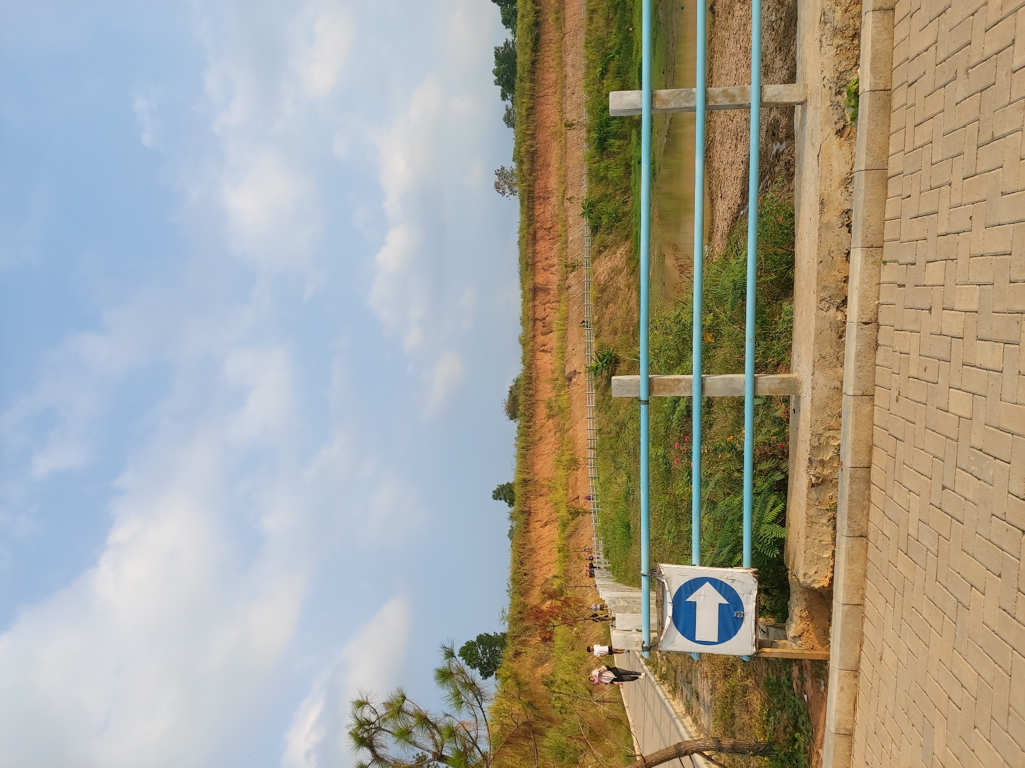 The image depicts an outdoor scene, likely a park or recreational area, with a paved walkway in the foreground and a body of water to the right, separated by a blue railing. A prominent white sign with a blue arrow pointing to the right is affixed to a post near the railing. In the background, a steep, eroded embankment is visible, covered in dry grass and vegetation, with trees at the top. Several individuals are walking on a paved path that winds along the embankment. The sky is blue with scattered white clouds. The scene suggests a serene environment with people engaging in leisure activities. While the image is stated to be in Bandar Lampung, Indonesia, there are no specific landmarks or contextual elements within the image that definitively confirm this location.