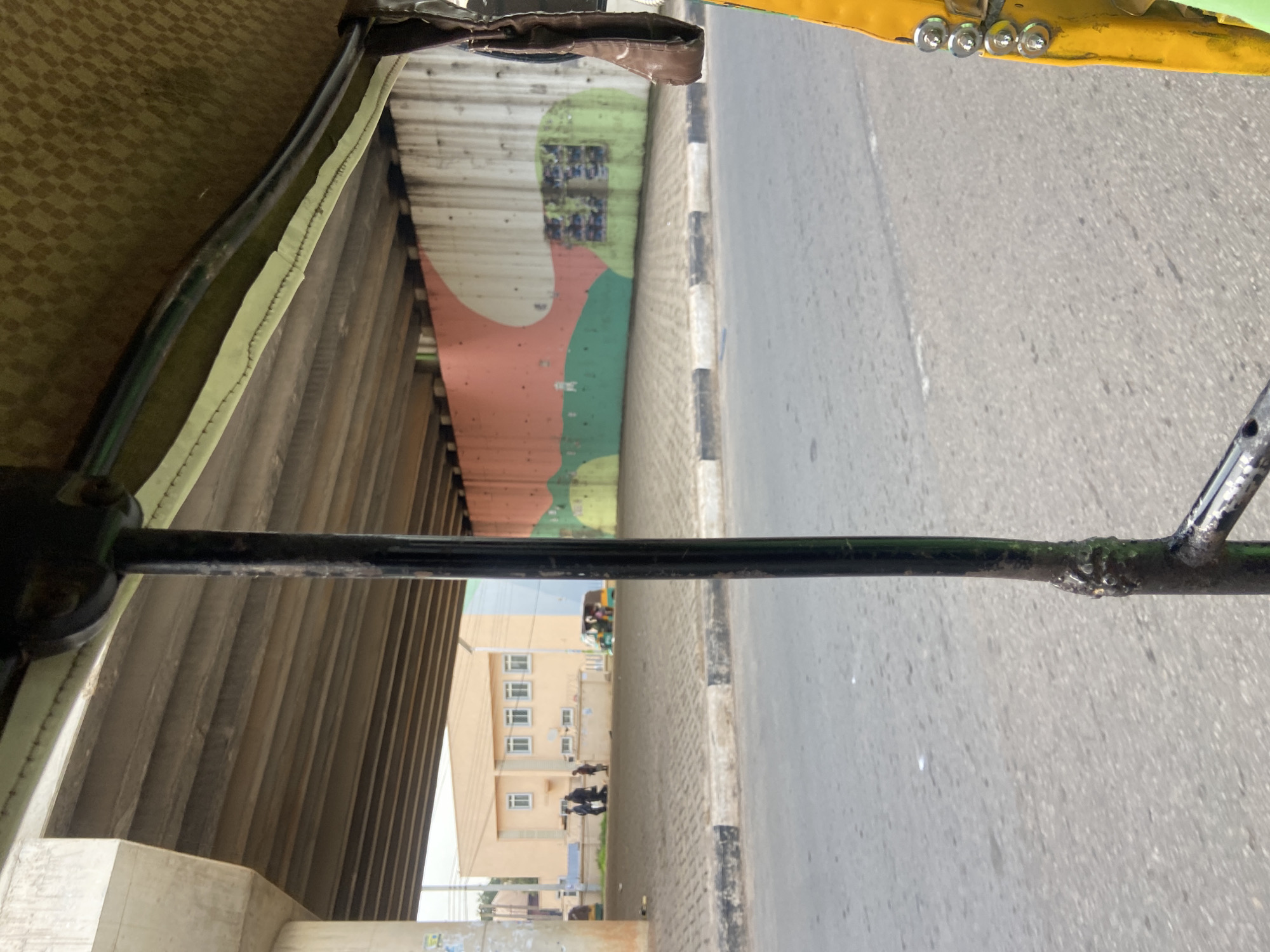 The image captures a scene in Jos, Nigeria, from the perspective of someone inside a vehicle, possibly a rickshaw or tuk-tuk, as indicated by the presence of a metal bar in the foreground and the interior of the vehicle on the left.
The setting appears to be urban, with a road and a pedestrian walkway visible on the right. The walkway is bordered by a curb with black and white markings. Beyond the walkway, a building with multiple windows is visible. In the distance, another vehicle, likely a tuk-tuk, is also present. The wall under an overpass or bridge is decorated with abstract shapes in red, pink, and green.
The time of day seems to be daytime, as suggested by the bright lighting and the clear visibility of the surroundings. There are no strong indicators of the weather, but the lighting suggests it is not overcast.
There are a few people visible in the image. Some are walking on the walkway near the building, and others are inside the distant tuk-tuk. The overall mood is neutral, depicting everyday life.
There is no visible text in the image.