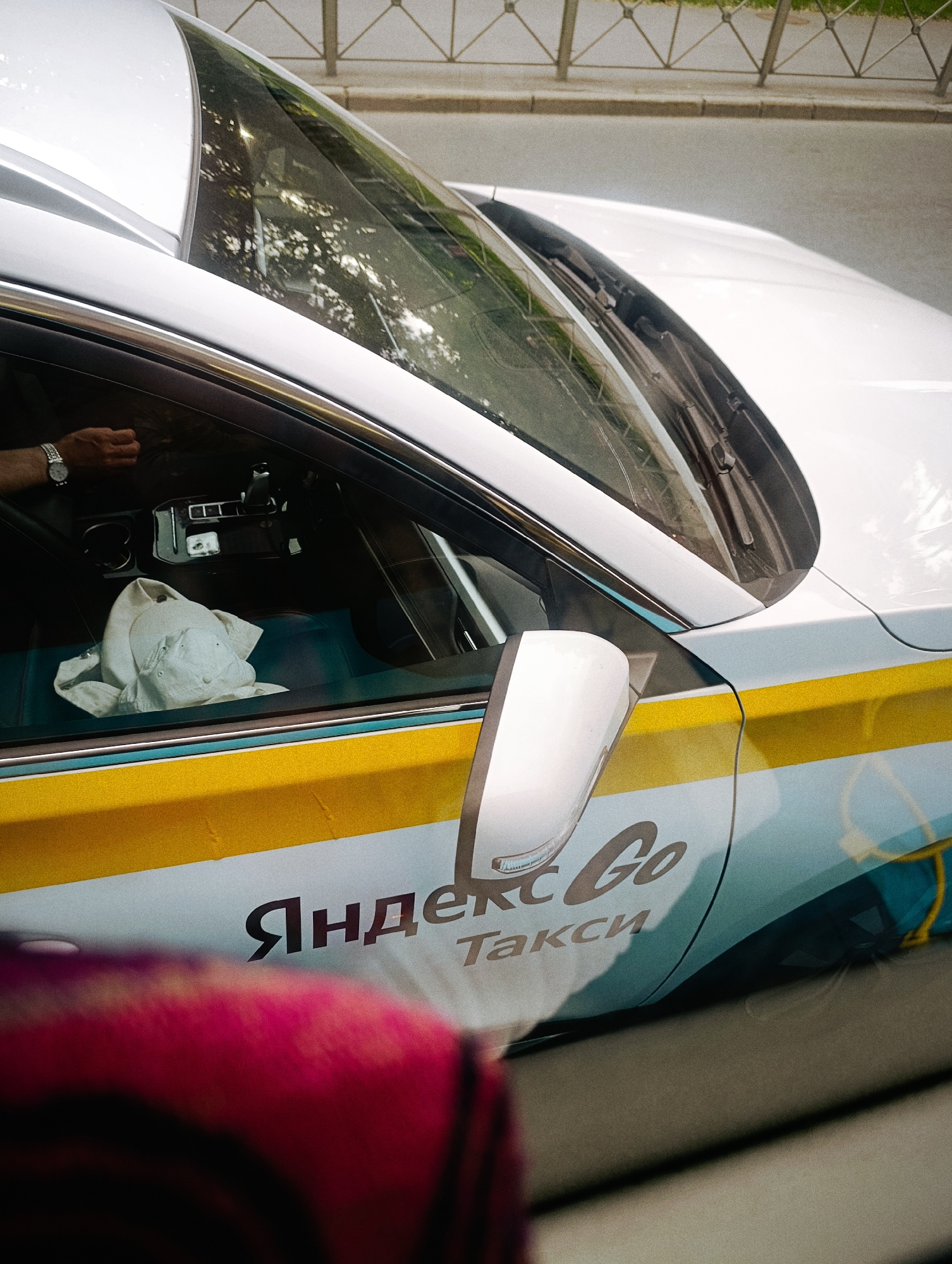 This media file shows a close-up of the side of a white car, likely a taxi, with yellow and light blue accents. The car has a prominent side mirror and part of the windshield is visible, reflecting trees and the sky. The car's door has the text "Яндекс Go Такси" printed on it, which translates to "Yandex Go Taxi."
Inside the car, a white cap is visible on the passenger seat. A person's hand, wearing a watch, is visible near the steering wheel, suggesting a driver. The scene appears to be taking place outdoors, possibly on a street, indicated by the road and a fence in the background. The lighting suggests it is daytime. No specific weather conditions are evident, but the reflections on the windshield imply it is not raining. The overall mood is neutral, focusing on the car and its branding.