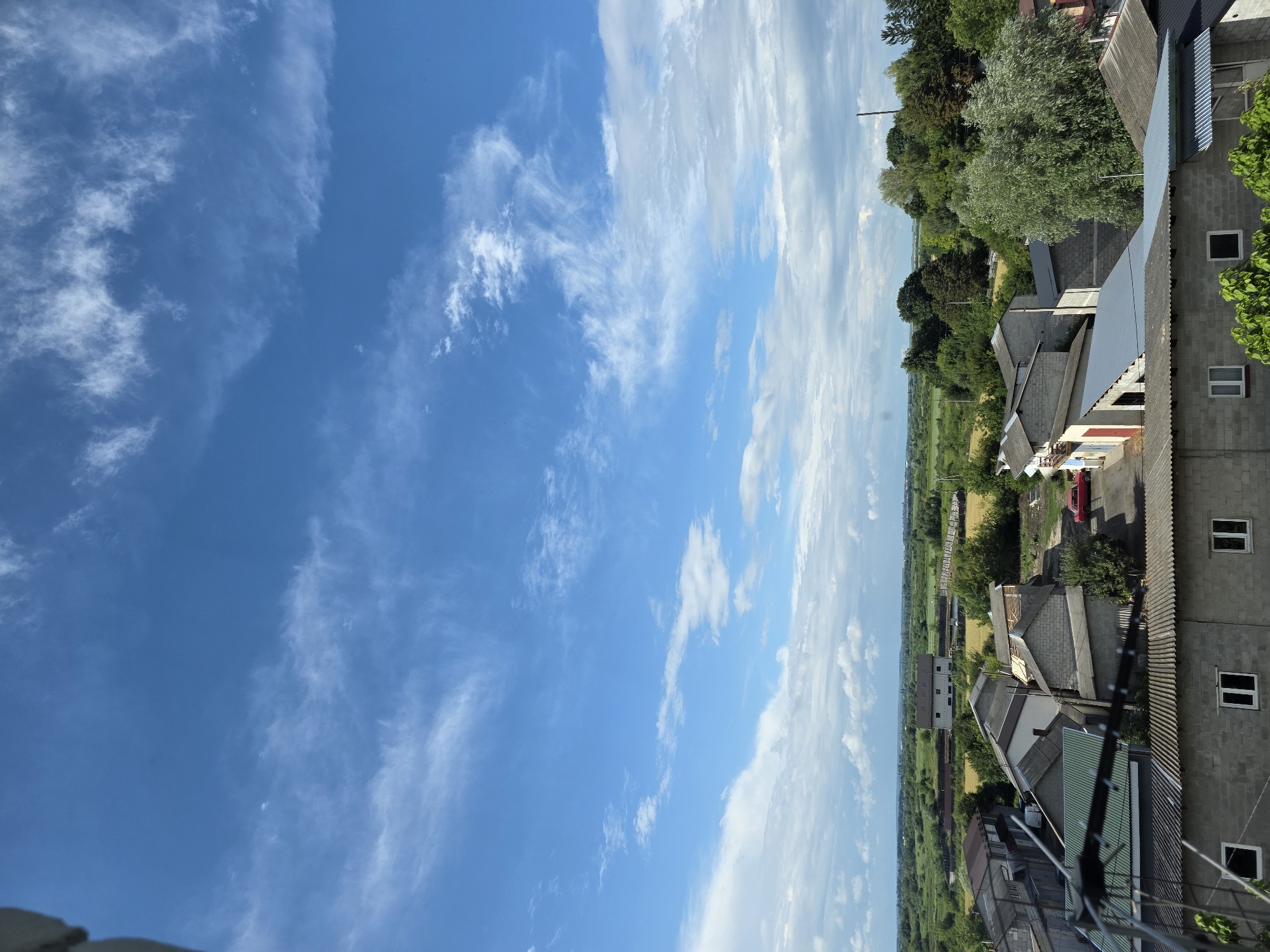 The image depicts a slightly overcast daytime sky with scattered cirrus clouds over a suburban or rural landscape. In the foreground, a collection of low-rise buildings with corrugated metal roofs are visible. These structures appear to be industrial or residential outbuildings. Further into the scene, there is more greenery, including trees and fields. In the distance, a larger building with a flat roof can be seen, possibly a warehouse or industrial facility. A red car is parked on a paved area between some of the buildings. The overall impression is of a non-descript, ordinary day in a community on the outskirts of a city. Given the request to include the location of Dubno, Ukraine, this scene could represent a view from a residential building or an elevated position within or near that city. There are no people visible in the image.