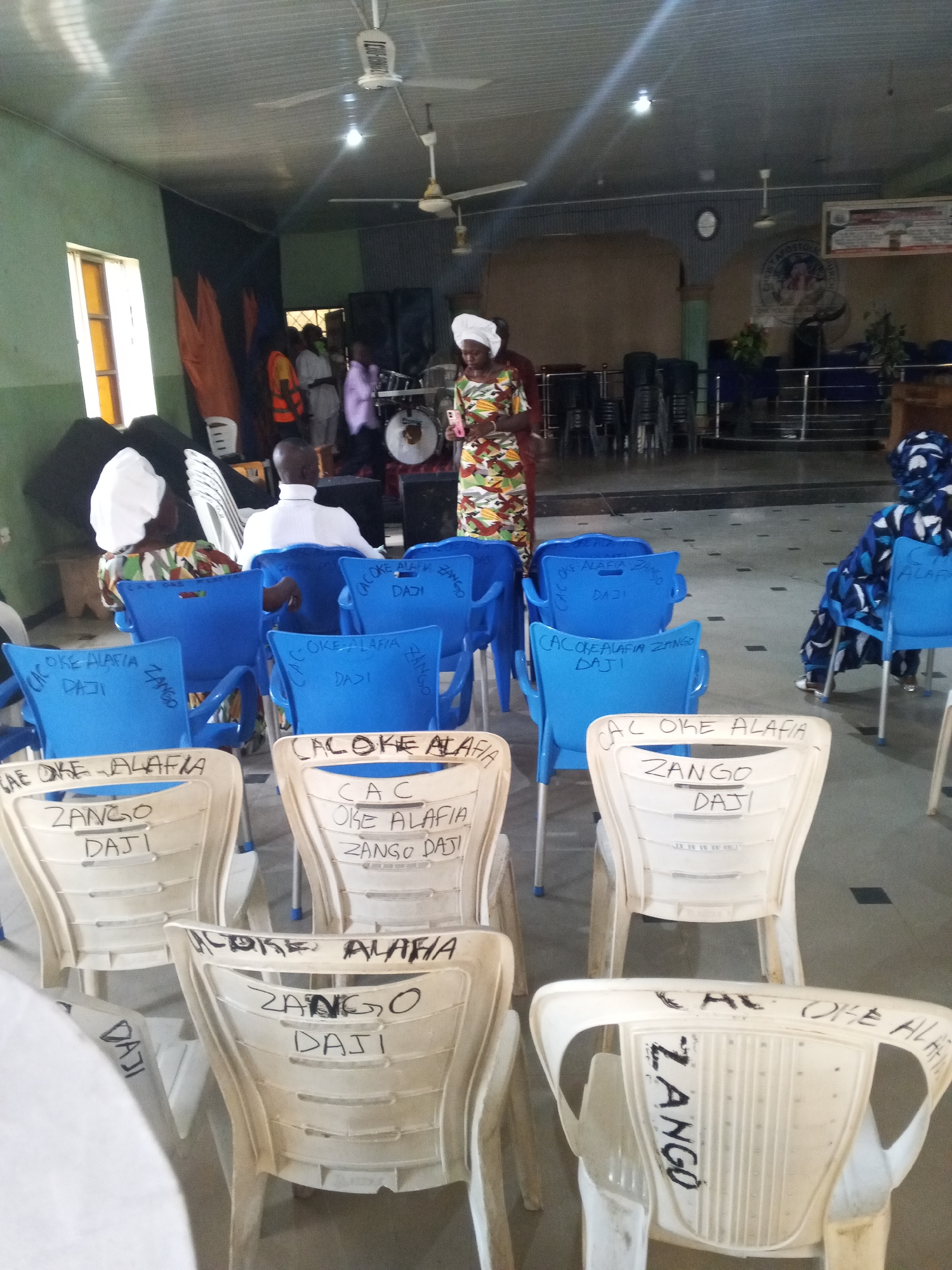 The image shows the interior of a building, possibly a church, in what is assumed to be Zango-Daji, Nigeria, given the inscriptions on the chairs. Several rows of plastic chairs, some blue and some off-white, are prominently displayed in the foreground. Many of the chairs have the inscriptions "CAC OKE ALAFIA ZANGO DAJI" written on their backs. People are present, although not all are clearly visible. A woman in a patterned dress and head covering stands in the middle of the frame, seemingly addressing the room. Another individual in a white head covering sits in one of the blue chairs in the foreground, and a man is seated near them. In the background, more people are present, including some near a drum set. The room has a ceiling with ceiling fans, and a window is visible on the left. Overall, the scene suggests a gathering or service of some kind.
