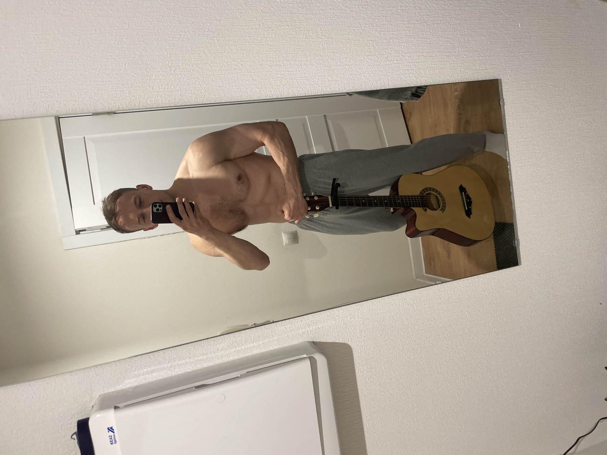 This mirror selfie captures a man standing, appearing shirtless and wearing grey sweatpants and white socks. He is holding a dark-colored smartphone, used to take the photo, which partially obscures his face. In his right hand, he holds an acoustic guitar, to which a capo is attached near the headstock. The man has short, dark hair and a visibly fit physique.
The reflection in the large, rectangular mirror shows an indoor room. The walls are light-colored, possibly white or off-white, with a subtle textured finish. A white door or door frame is visible in the background, and wooden flooring can be seen on the right side of the reflection.
Outside the mirror's frame, in the lower left foreground, a portion of a white object or appliance is visible, featuring blue text that reads "KEAZ Optima". A black cable lies on the floor near the bottom right edge of the mirrored surface. The lighting is internal, providing no specific time of day cues. The image presents a casual, personal moment within what appears to be a residence in Saint Petersburg, Russia.