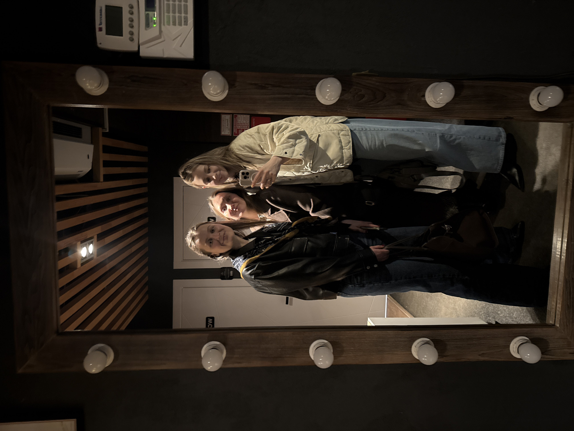 Three women are taking a selfie in a mirror with light bulbs around the frame. The woman on top is holding the phone. The woman in the middle is wearing a dark coat. The woman on the bottom is wearing a leather jacket. In the background, there are signs for the women's restroom and wooden slats on the wall. There is a security panel on the wall above the mirror. The location may be in Chaikovsky, Russia.