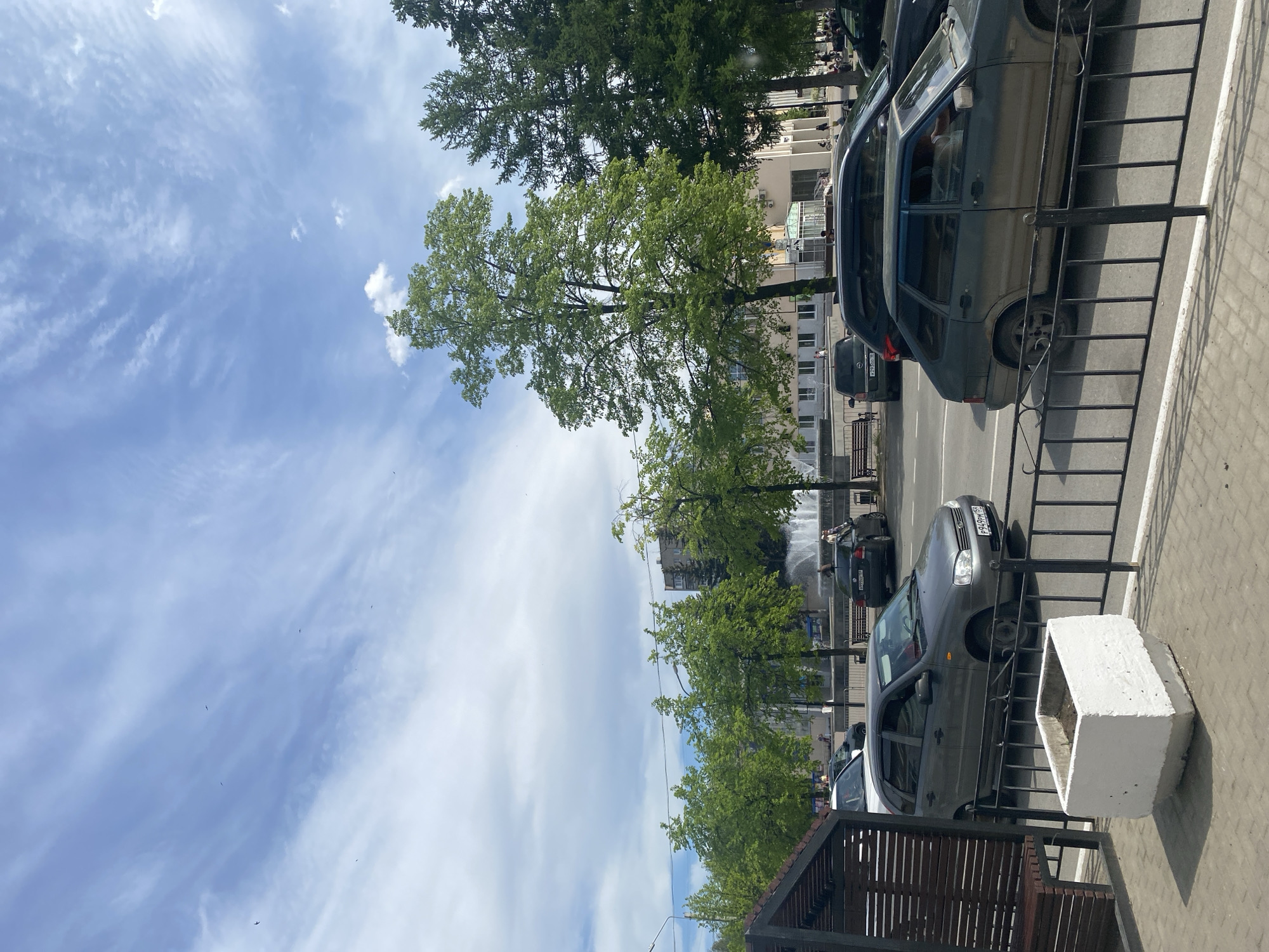 The image shows a street in Chaikovsky, Russia. There are several cars parked along the side of the road, next to a metal fence and white concrete planters. Trees line the street, and in the background, there appears to be a fountain and several buildings. The sky is blue with white clouds.