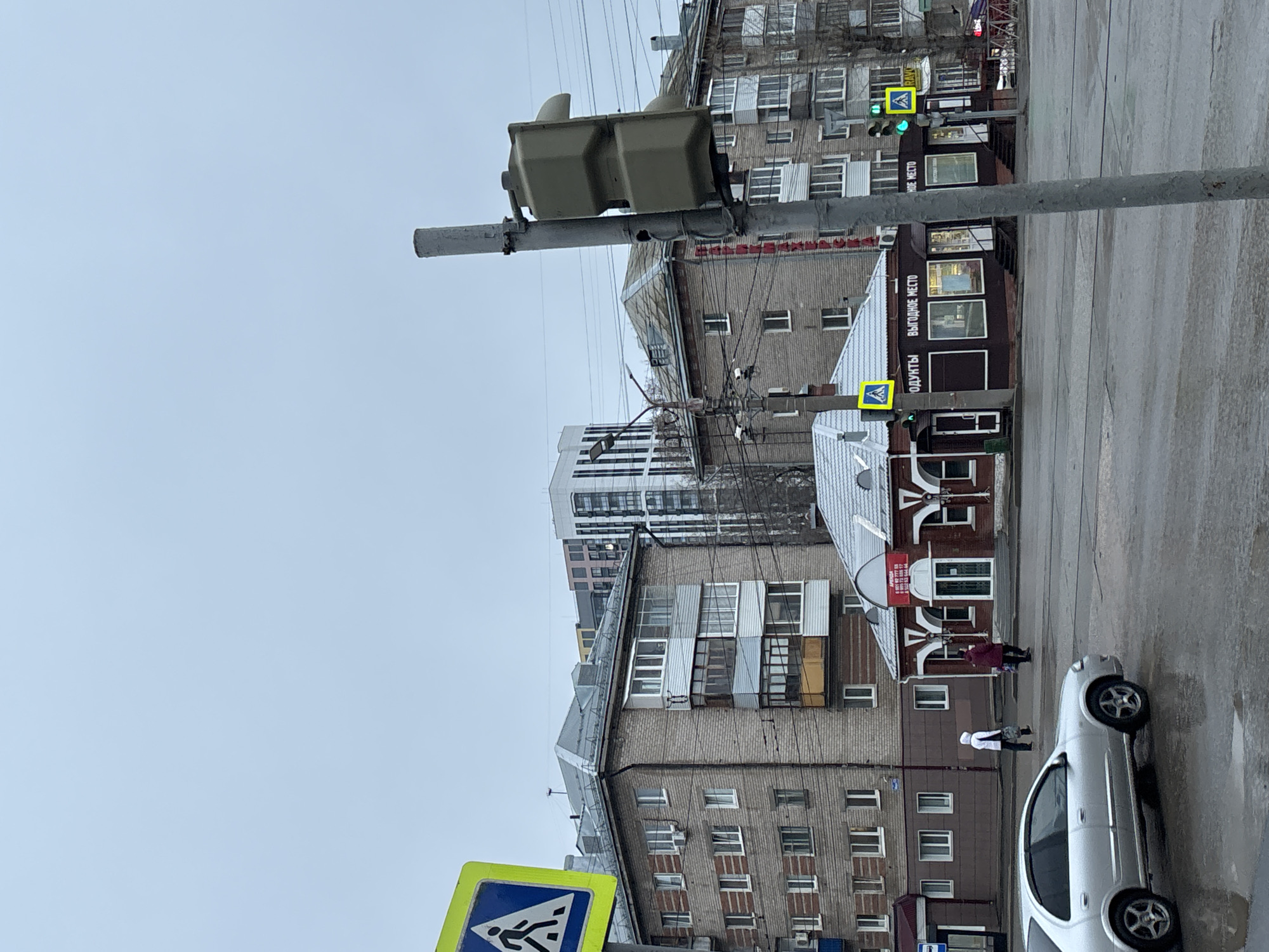 The image shows a street scene in Perm, Russia. The sky is overcast and grey. In the foreground, a traffic light pole and a pedestrian crossing sign are visible. A silver car is parked on the side of the street. People are walking on the sidewalk. Several buildings line the street, including brick apartment buildings and a two-story wooden building with a sign in Russian. Power lines crisscross overhead. A traffic light is illuminated with a green signal.