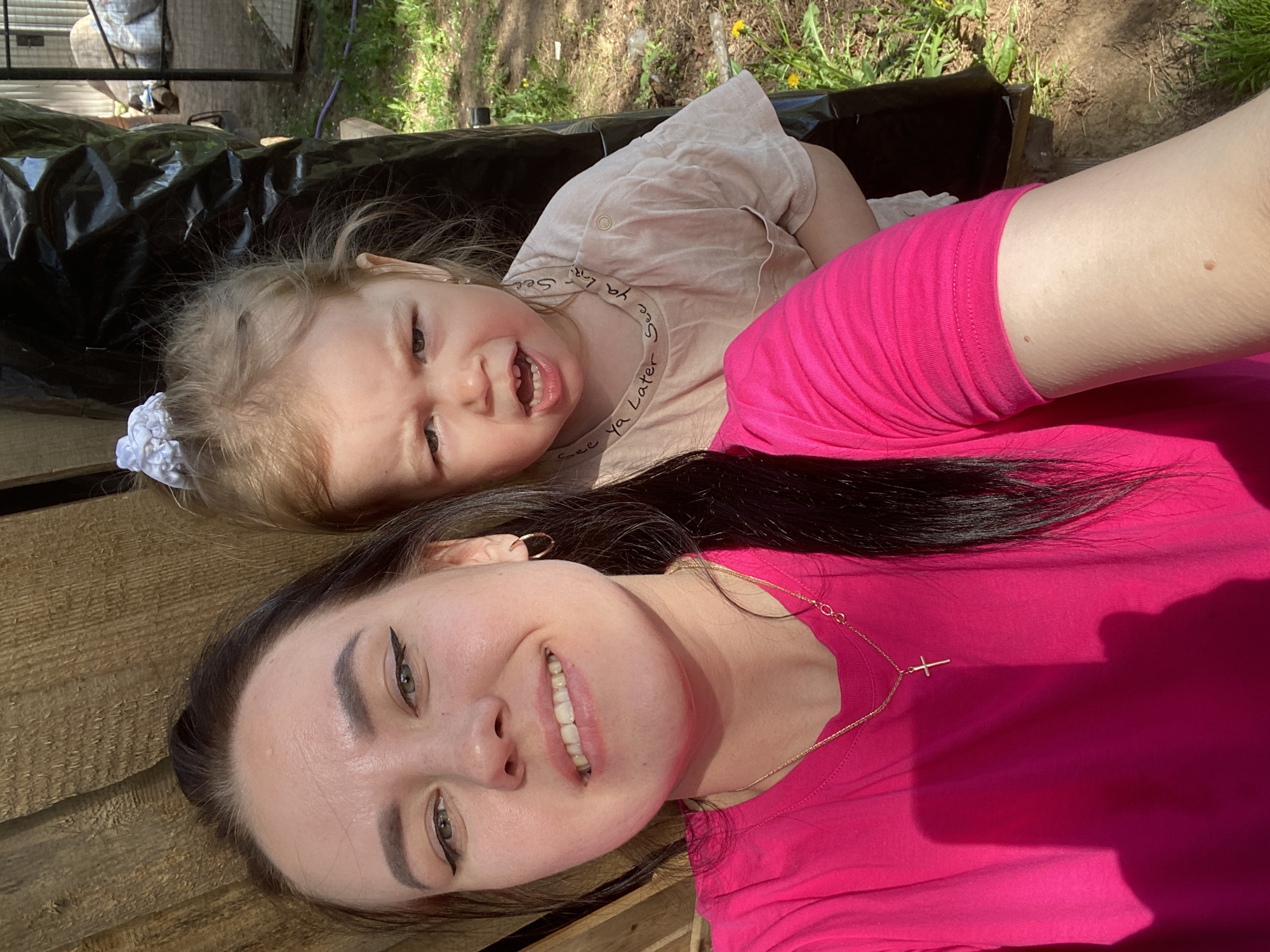 A selfie shows a woman and a young child lying down outdoors. The woman has dark hair pulled back, fair skin, and is wearing a pink t-shirt and a gold necklace with a cross pendant. She is smiling at the camera. The child, who appears to be about two years old, has light hair and is wearing a light-colored shirt with text. The child is grimacing at the camera, showing their teeth. They are lying on what appears to be wooden planks. Behind them are items covered with black plastic. Green vegetation and dandelions are visible in the background. The location is outdoors in Chaikovsky, Russia.
