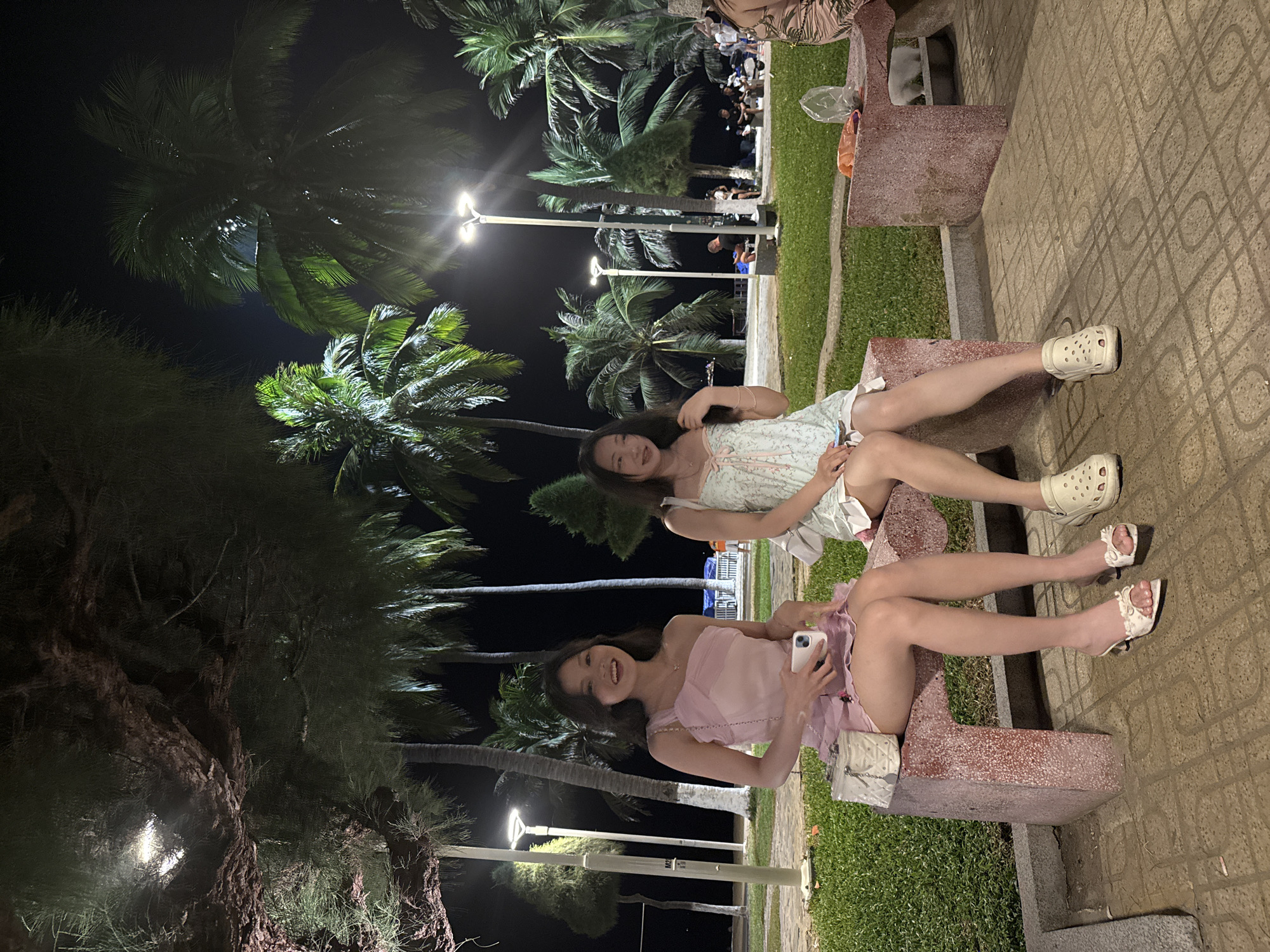 Two young women sit on a stone bench. One wears a pale pink dress and white heels, holding a smartphone. The other wears a light patterned dress and white Crocs. They are outdoors at night, with palm trees and street lights visible. The ground is paved with a patterned design. The location is Nha Trang Ward, Vietnam.