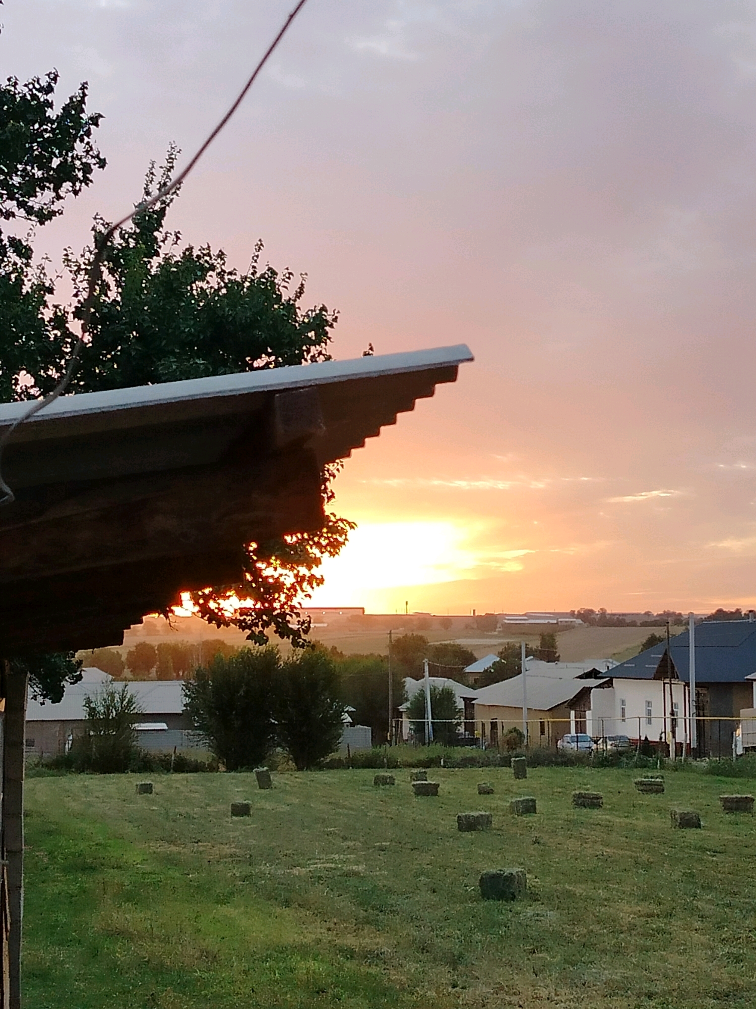 This image captures a rural landscape at sunset. In the foreground, a portion of a wooden structure with a corrugated roof juts into the frame from the left, casting a shadow. Beyond this structure, a green field is dotted with numerous rectangular hay bales. In the middle ground, a cluster of trees and several houses with pitched roofs are visible. A light-colored car is parked in front of one of the houses. The background features more trees and distant buildings, with a power line crossing the frame. The sky is painted with hues of orange, pink, and yellow, indicating the setting sun, which is partially obscured by clouds and distant trees. The scene appears to be located in the village of Kölkent, Kazakhstan.