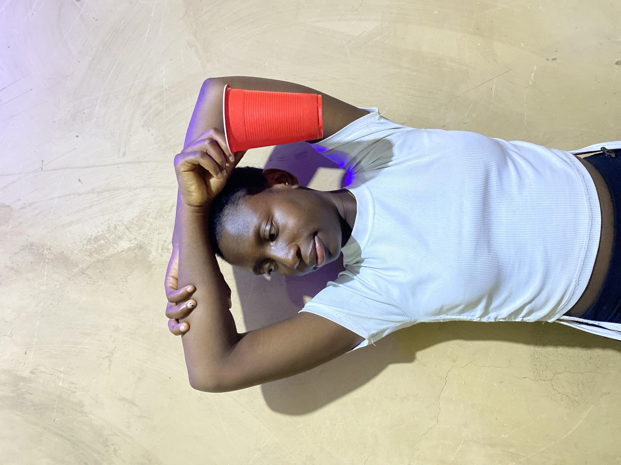 A young person with dark skin and short dark hair is depicted lying horizontally on a light-colored, textured surface, likely a floor or ground, in Kuru Dungus, Nigeria. They are wearing a white ribbed short-sleeved top and dark bottoms, with only the waistband visible.
The subject's right arm is bent and extended above their head, holding a bright red plastic cup. Their left arm is also bent, with the hand resting near their shoulder. The person's face is turned towards the viewer, looking downwards and to the right with a relaxed, slightly smiling expression, suggesting contentment or playfulness.
A subtle purple and blue light illuminates the upper left portion of the image, casting a glow on the subject's left arm and hair, indicating an artificial light source. The background surface shows faint lines and scratches, adding to its textured appearance. The scene conveys a casual and calm moment, possibly taking place indoors or in a sheltered area during the evening or night. No text is visible in the image.