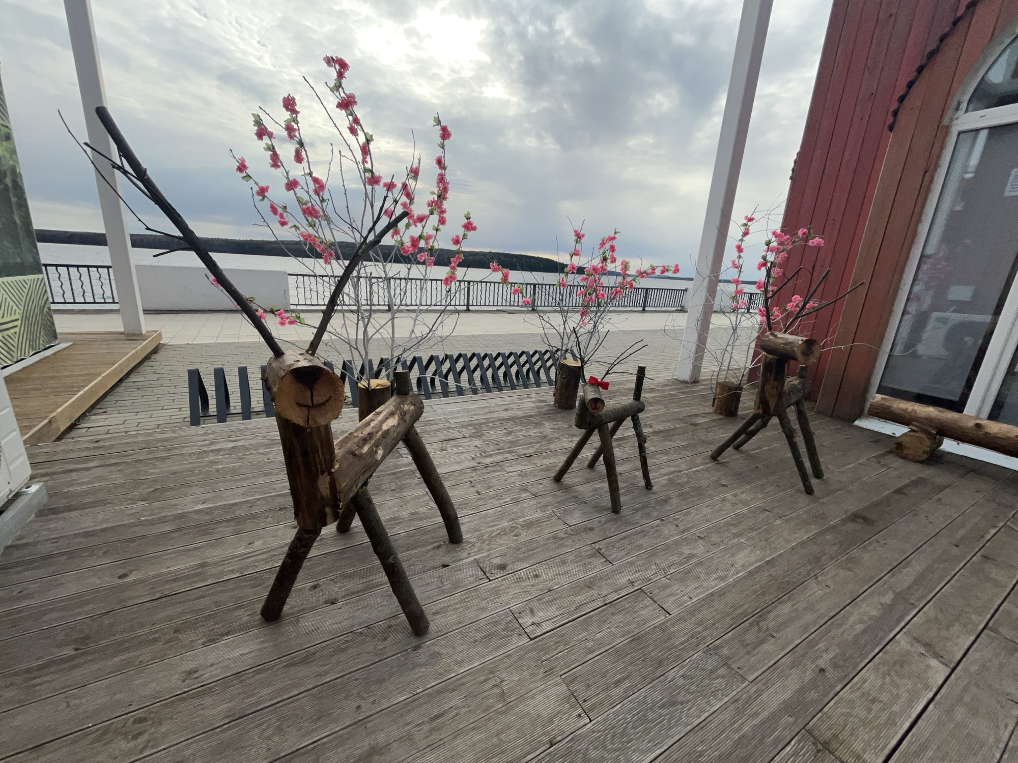 On a wooden deck in Chaikovsky, Russia, three handcrafted wooden deer sculptures stand on a cloudy day. The largest deer is in the foreground, constructed of thick, rough-hewn logs with stick antlers. A smaller deer with a red bow tie stands behind it, and the smallest is near a tilted, reddish-brown wooden structure with a window. Each deer is adorned with thin, leafless branches and delicate pink blossoms. A metal railing borders a body of water in the background. The cloudy sky adds to the scene's serene and rustic ambiance.