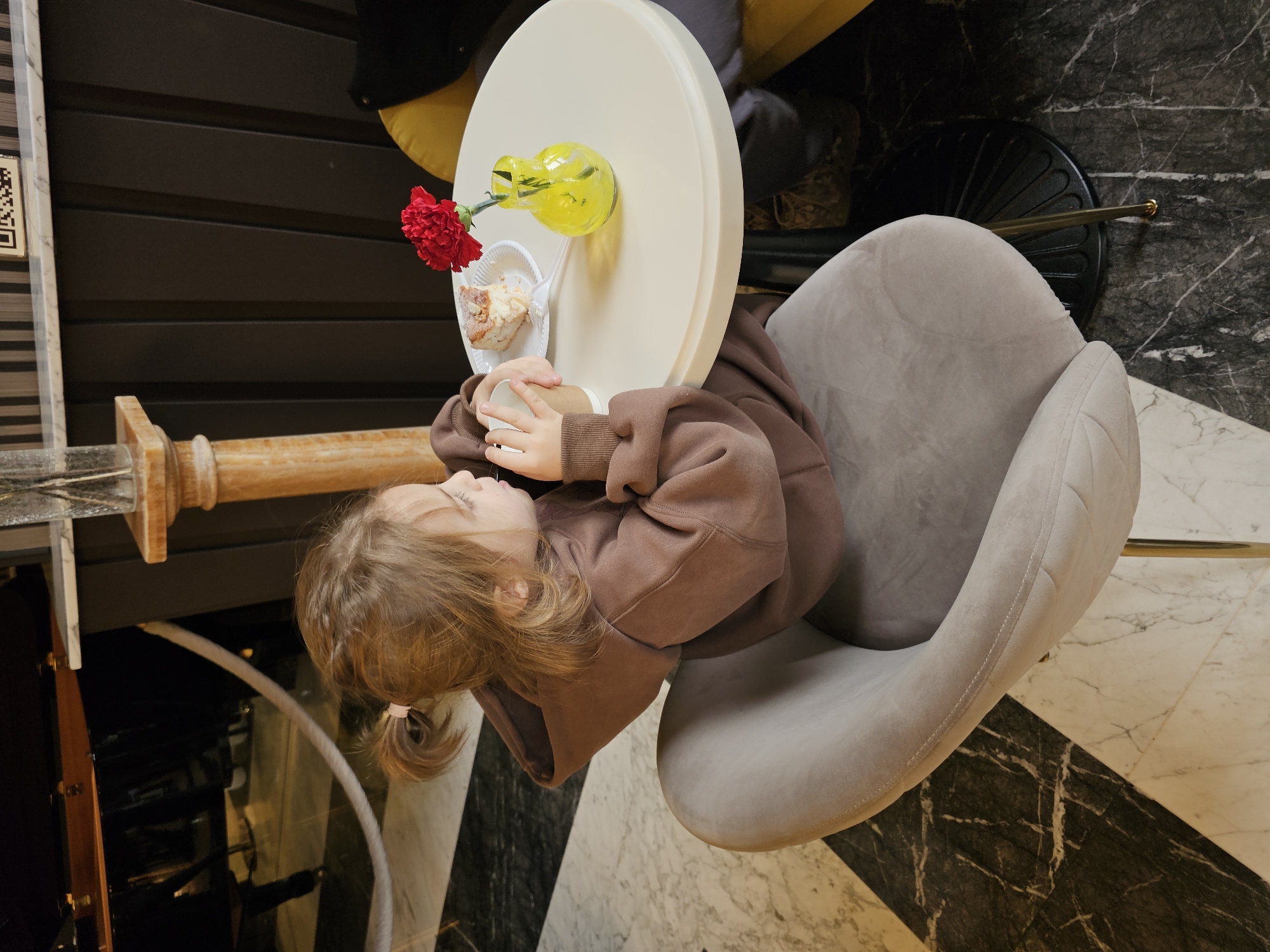 A small child wearing a brown hoodie sits in a grey velvet chair at a round, white table. On the table is a piece of cake on a white plate, a small yellow vase with a single red flower, and a paper cup. The floor is tiled with a marble pattern. The scene is located in Архангельское, Russia.