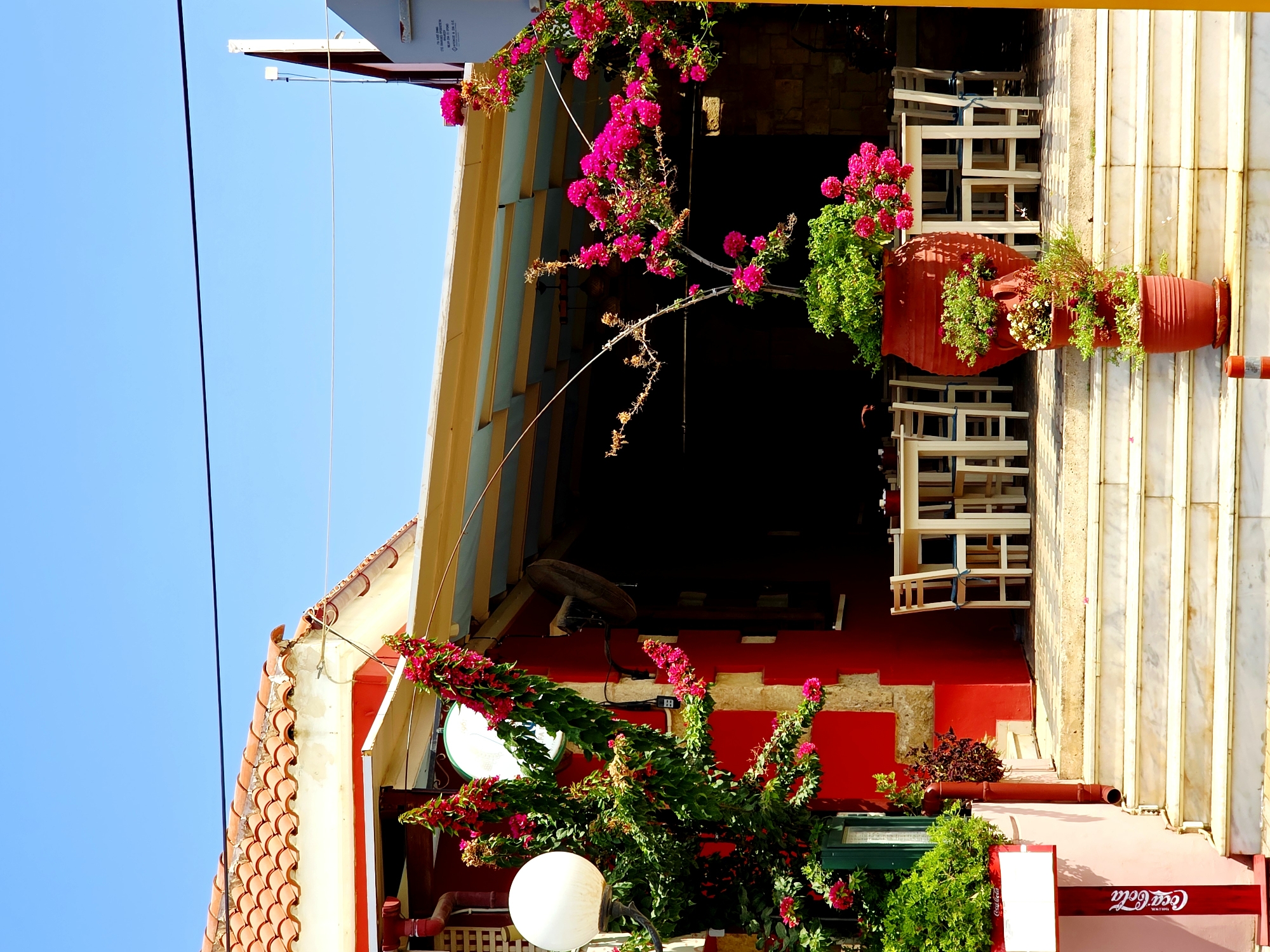 The image depicts the exterior of a vibrant red building in Daratsos, Greece, likely a restaurant or cafe, bathed in bright sunlight under a clear blue sky. The building has a traditional tiled roof and a yellow awning extending over an outdoor seating area. In front of the building, a set of white marble stairs leads up to the entrance.
Lush green plants and bright pink bougainvillea cascade from planters and hang from trellises, adding a burst of color to the scene. Several tables and chairs are visible under the awning, set for dining. A large, round white lamppost stands to the left, adorned with more flowering vines. A Coca-Cola branded stand is also present near the entrance. The overall impression is one of a charming and inviting establishment in a Mediterranean setting.