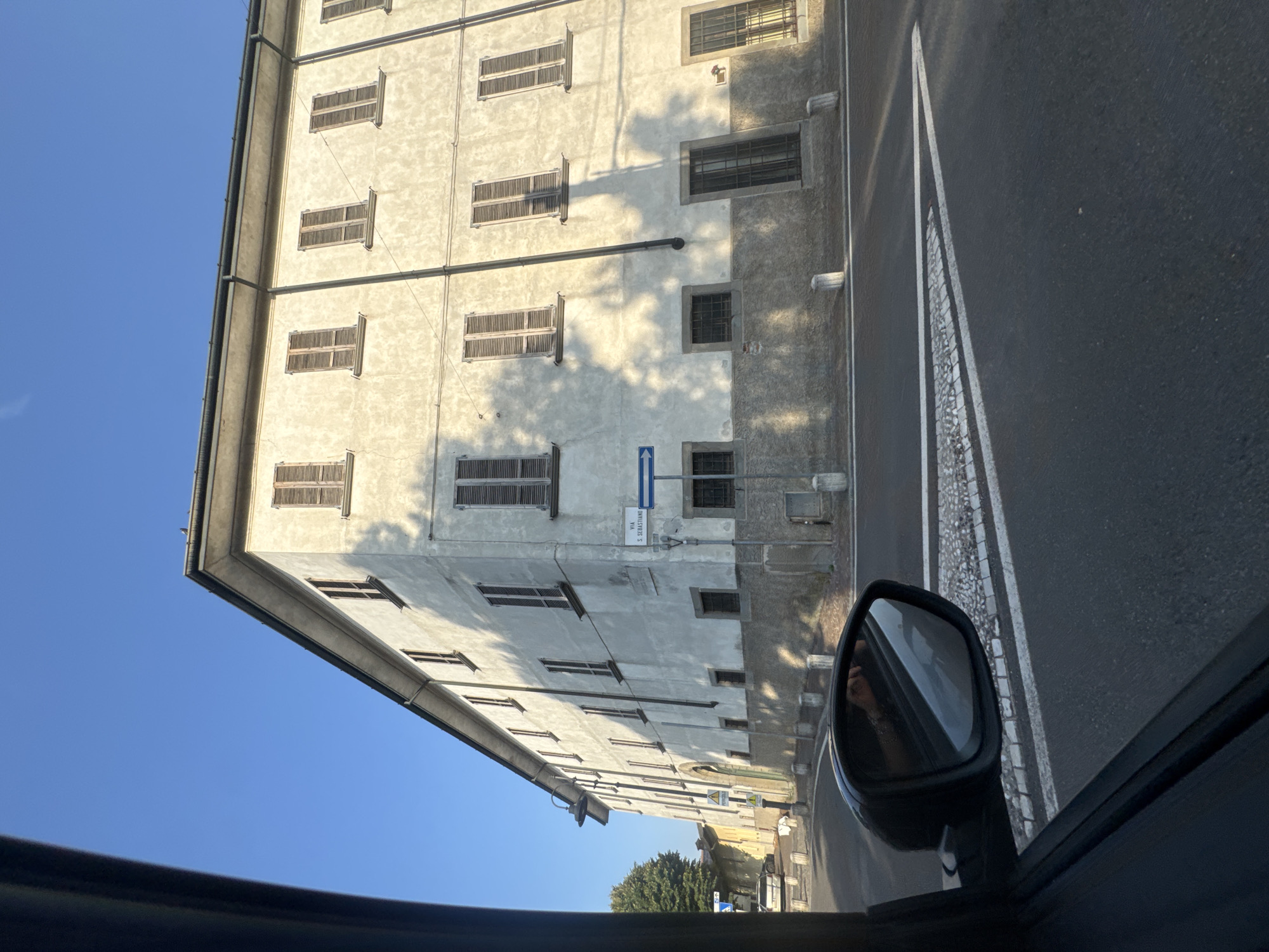 The image shows a view from inside a car, looking out at a building and a street in Chiari, Italy. The building is a multi-story structure with a textured, light-colored facade and numerous windows. Many of the windows have wooden shutters. The building appears to be residential or office space.
The street in front of the building is paved with asphalt and has white lane markings. There is a cobblestone border along the side of the street. A car's side mirror is visible in the lower right corner of the image, reflecting a person's hand and arm, likely the driver.
A street sign is visible on the building, with text that reads "VIA S. SEBASTIANO". There is also a blue directional sign with an arrow. The scene is bathed in sunlight, suggesting it is daytime, likely late afternoon due to the long shadows cast by the building and trees. The sky is clear and blue. The overall impression is a quiet street scene in an Italian town.