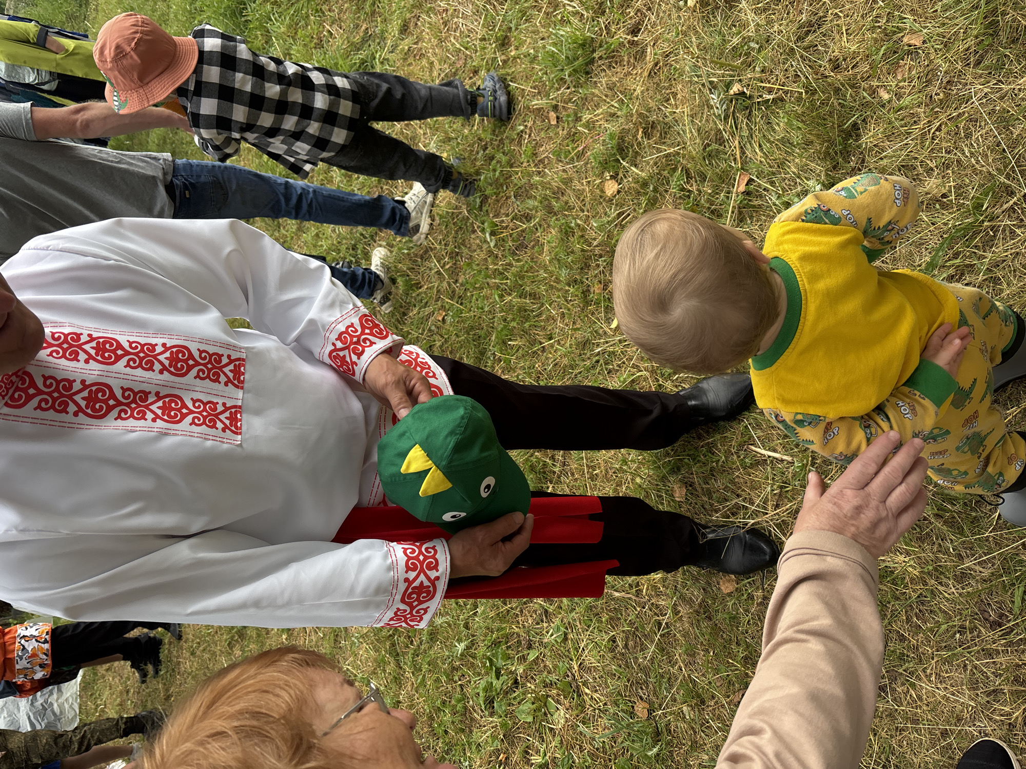 The image captures a moment outdoors on a grassy area, possibly in a park or rural setting. In the foreground, a person wearing a beige sleeve is extending their hand towards a toddler. The toddler, facing away from the camera, is wearing a yellow long-sleeved shirt with green trim and yellow pants with a dinosaur pattern. The toddler is looking down at the grass.
Behind the toddler, a person in a white shirt with red embroidery and a red sash is holding a green baseball cap with yellow dragon-like spikes and cartoon eyes. To the right of this person, another individual is visible, wearing a plaid shirt and an orange hat. The background shows hints of other people and a green landscape. The overall impression is a casual gathering or event.