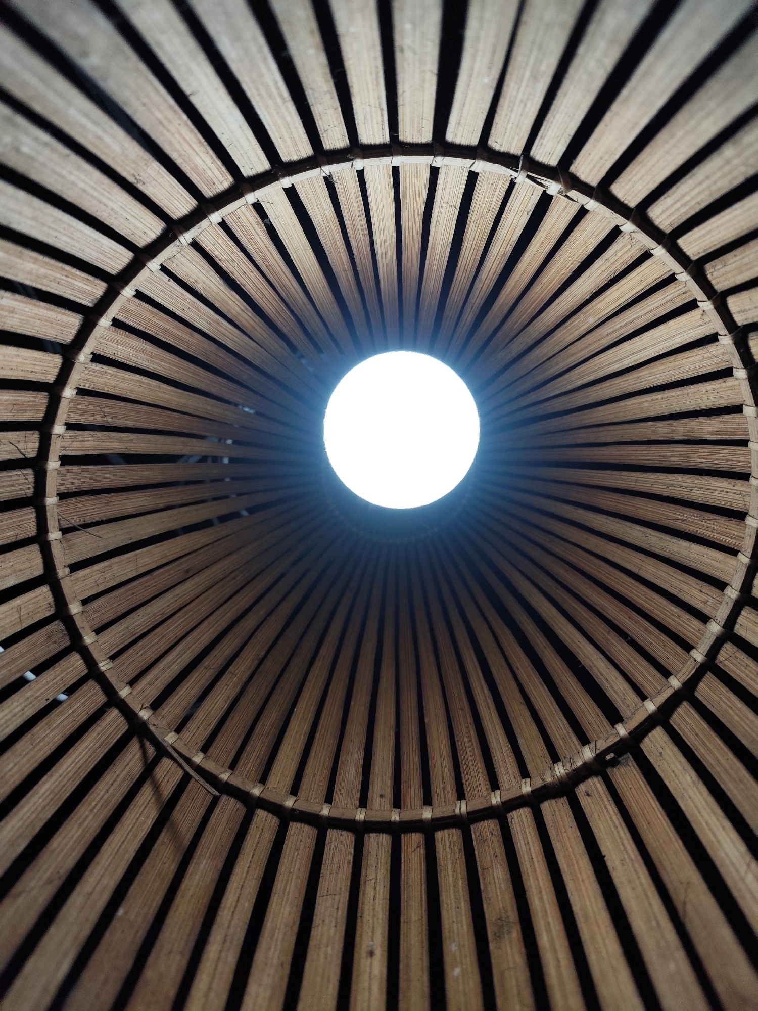The image shows an upward view through a woven bamboo structure, likely a lampshade or decorative element. The bamboo strips radiate outwards from a central circular opening, where a bright light source is visible. The texture of the bamboo is evident, and the overall tone is warm due to the color of the bamboo and the light. The location is presumed to be Da Lat, Vietnam, based on the prompt but no information in the image confirms the location.